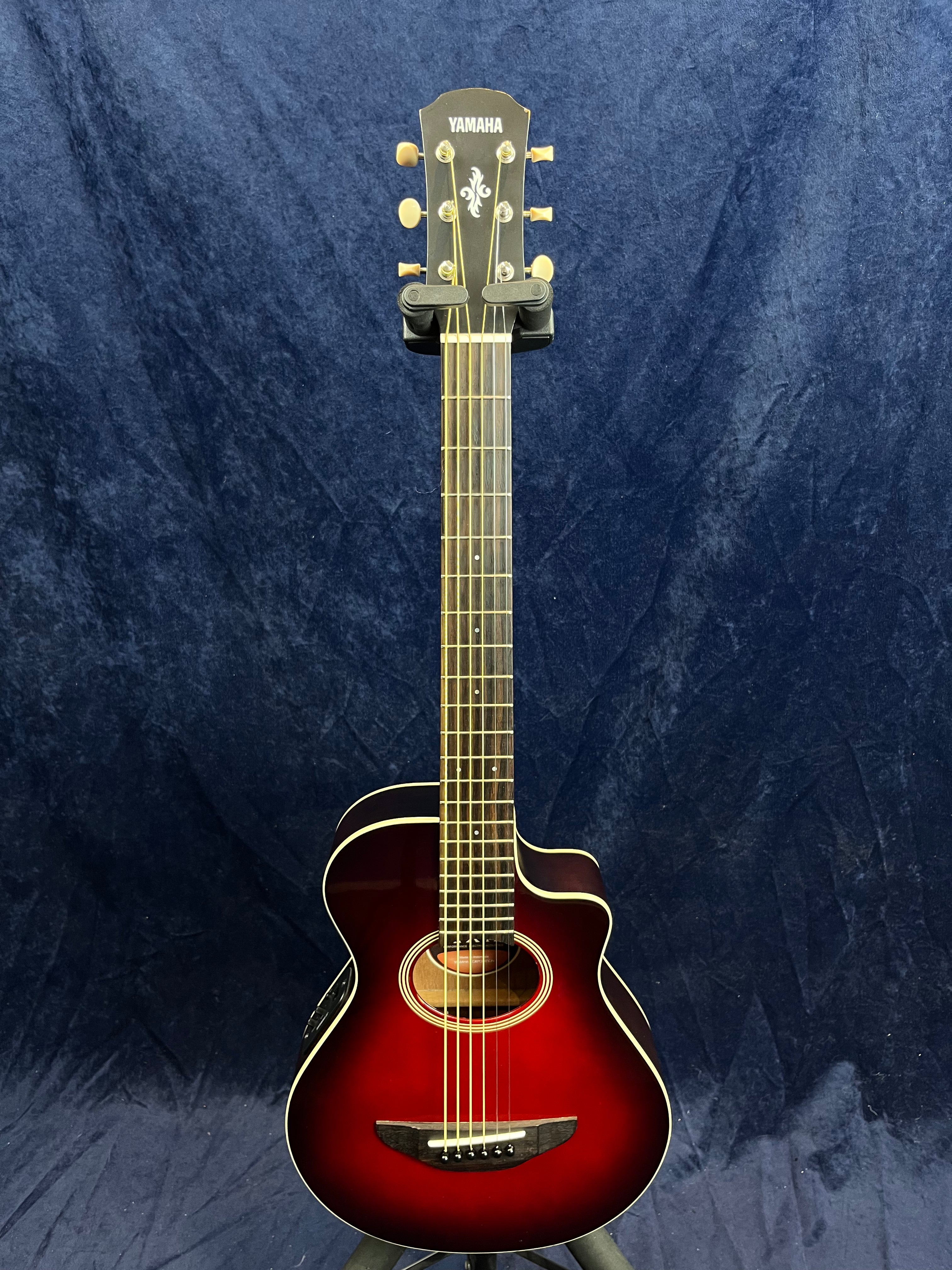 Yamaha acoustic guitar with a glossy red sunburst finish, cutaway body, six strings, and gold tuning pegs, displayed upright against a dark blue backdrop.