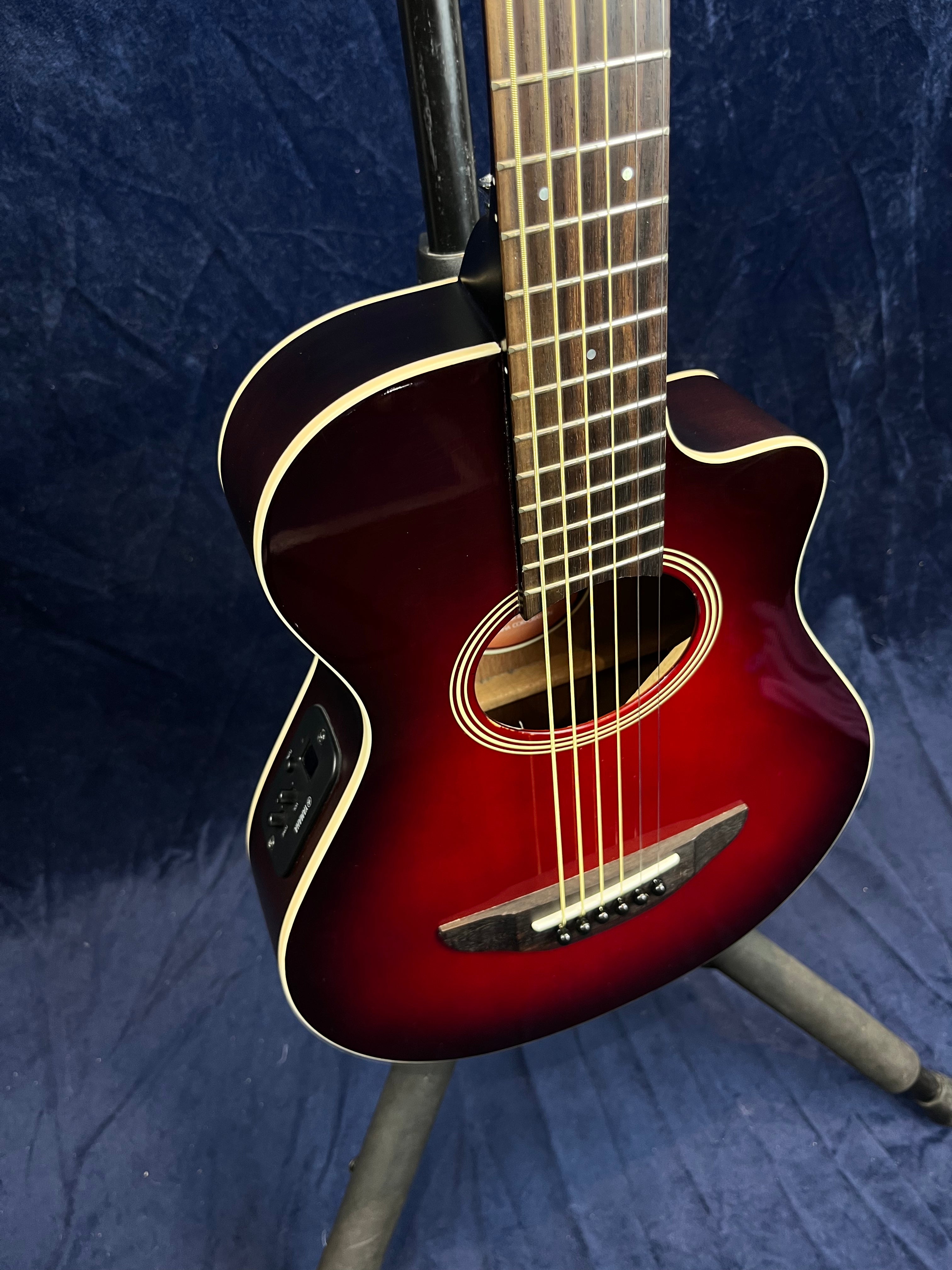 Acoustic guitar with a glossy red sunburst finish, cutaway body, electronic controls on the side, and cream binding on edges, resting on a stand.