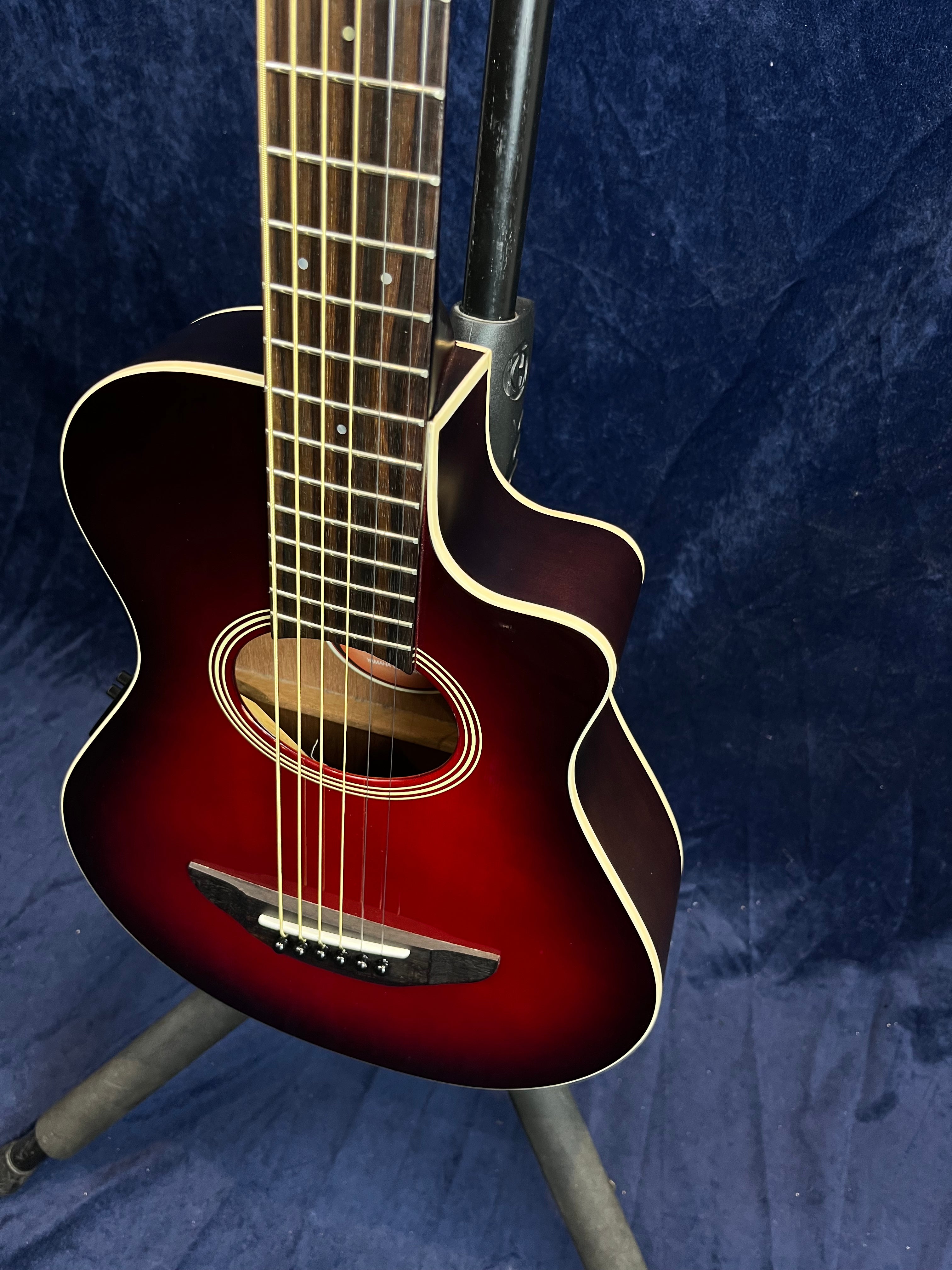 Red sunburst acoustic guitar with a cutaway body, cream binding, six strings, and a rosewood fingerboard, standing on a tripod stand.