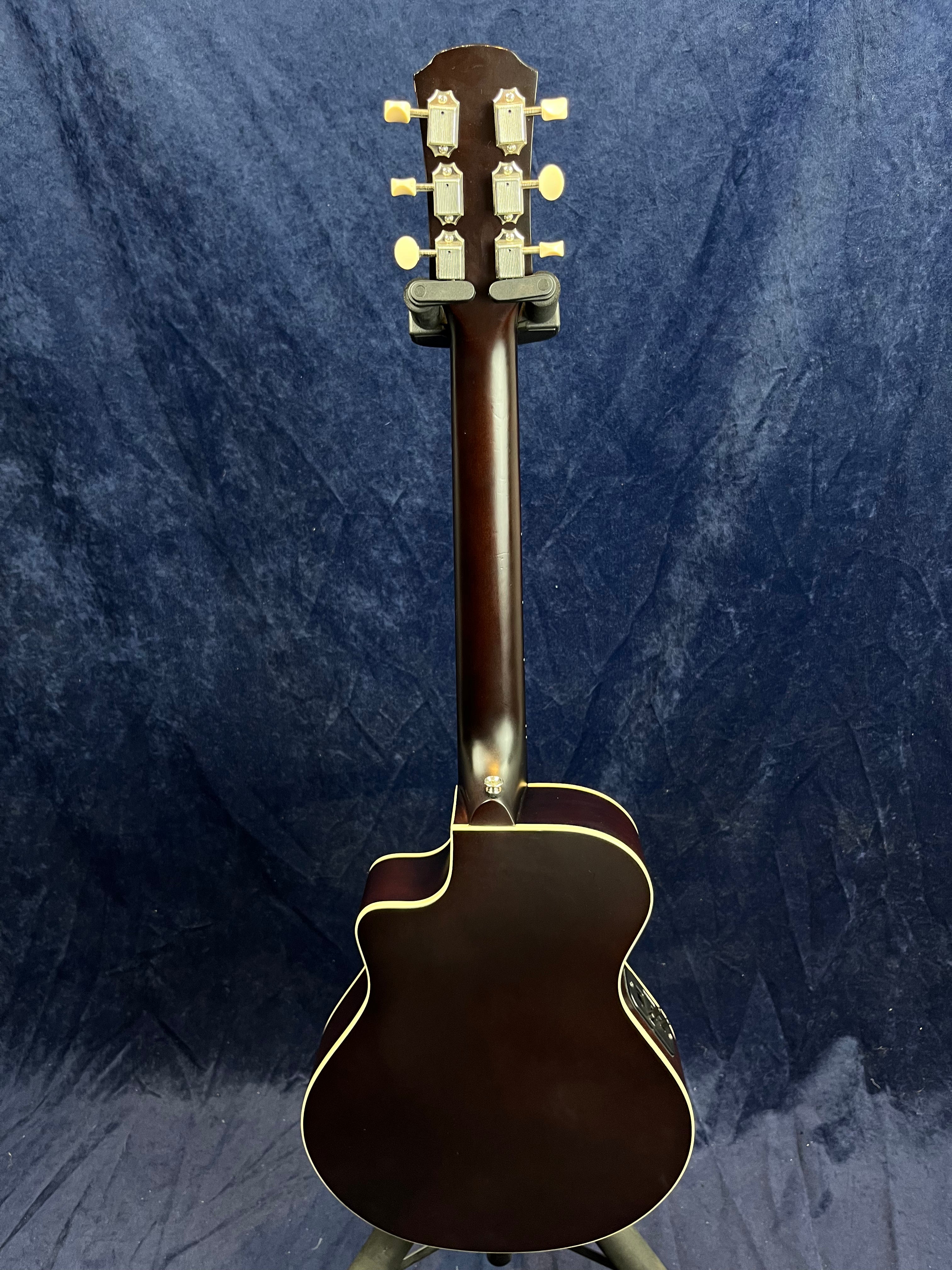 Dark brown acoustic guitar viewed from the back, showing tuning pegs, neck, and body with light-colored trim against a dark blue background.