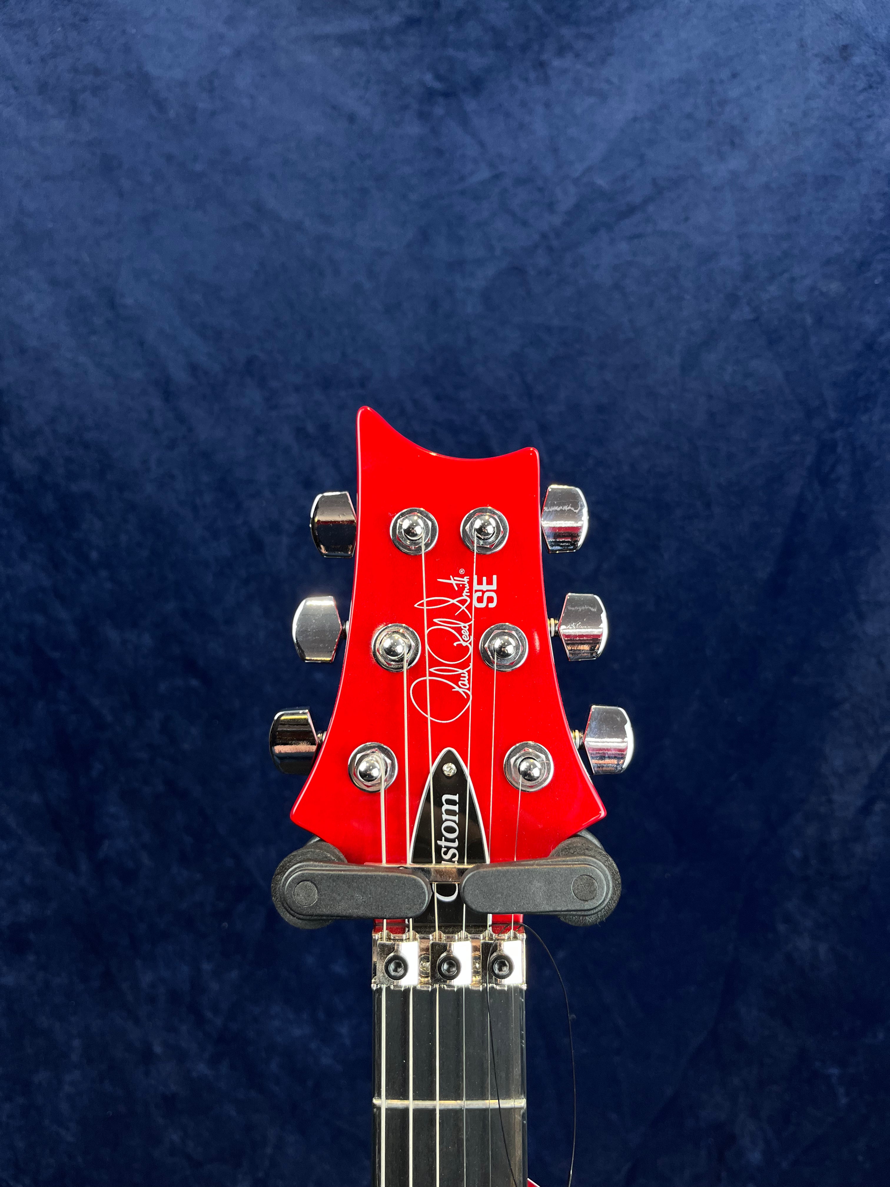 Red electric guitar headstock with six chrome tuning pegs and a signature logo, set against a dark blue background.