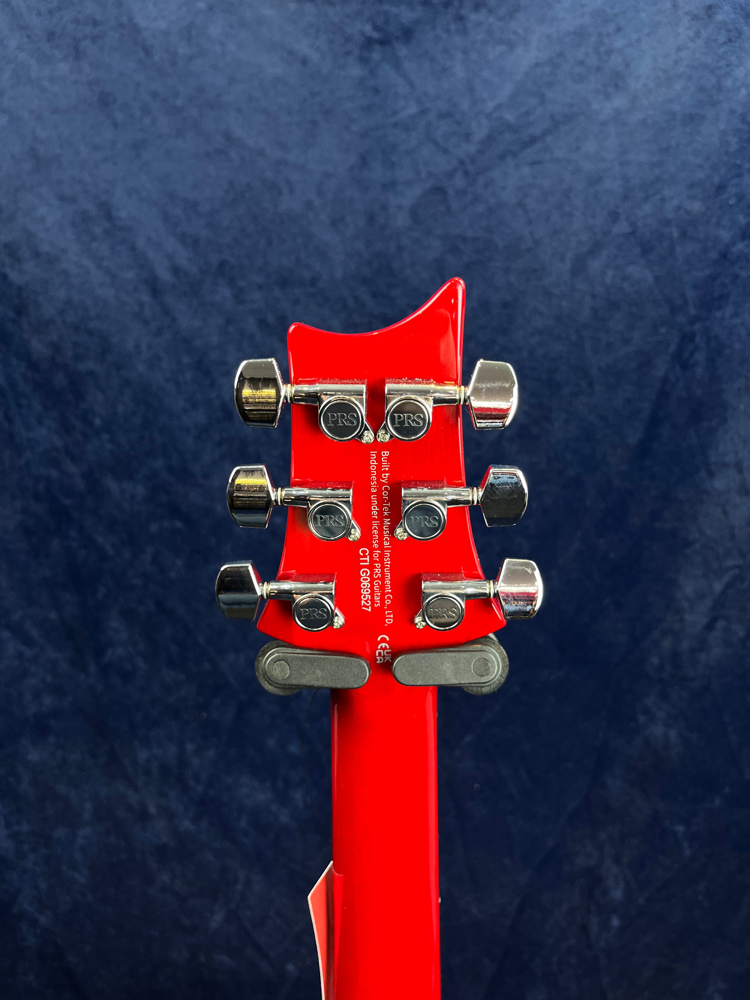 The back of a bright red electric guitar headstock with six chrome PRS tuning pegs and white serial number text against a dark blue background.
