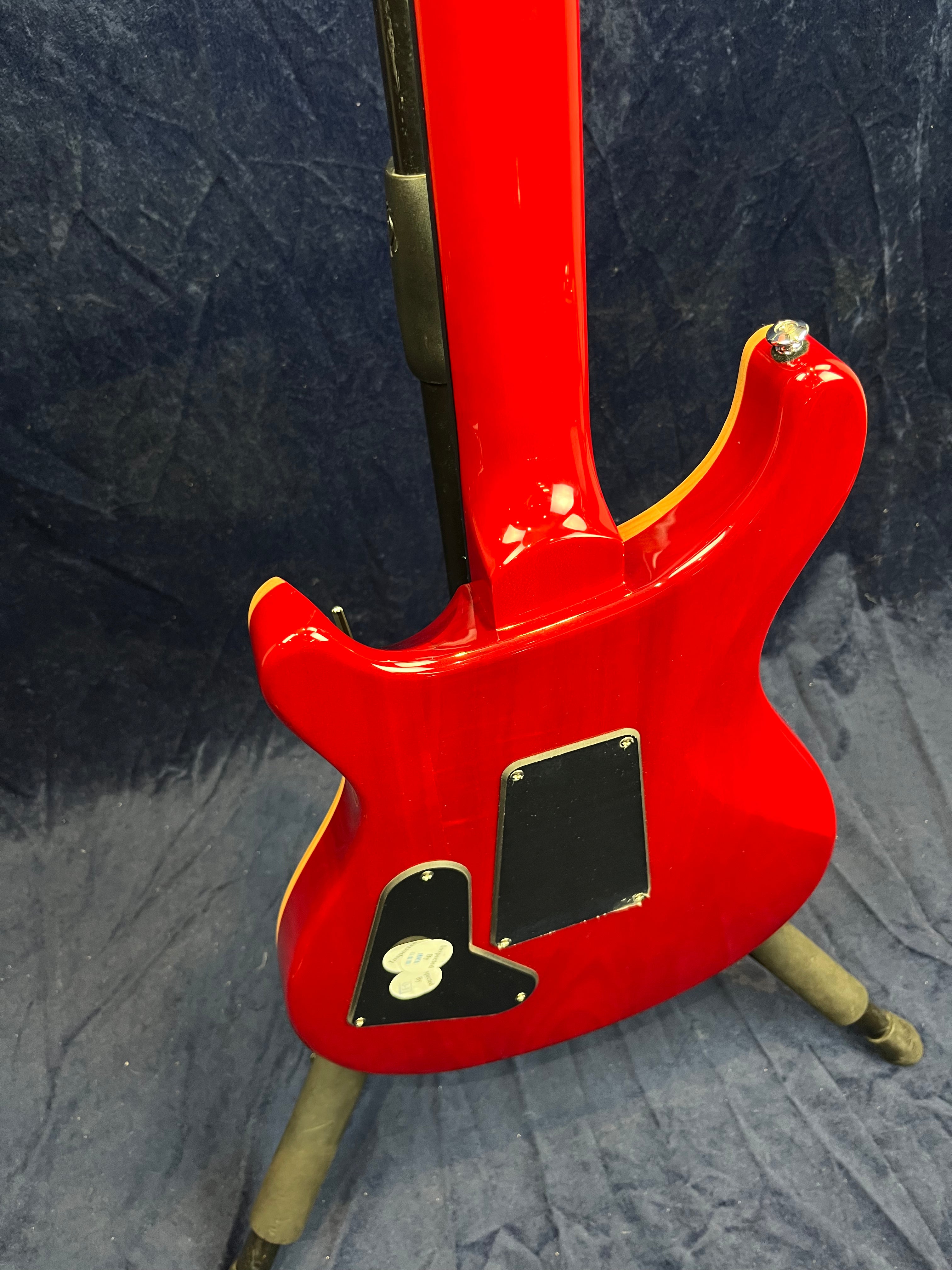 Bright red electric guitar viewed from the back, showing the neck joint, two black plastic back plates, and yellow edge binding, resting on a stand.