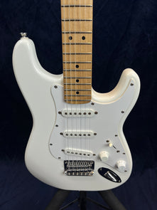 White electric guitar with three single-coil pickups, two control knobs, a five-way switch, maple neck, and a chrome bridge against a dark background.