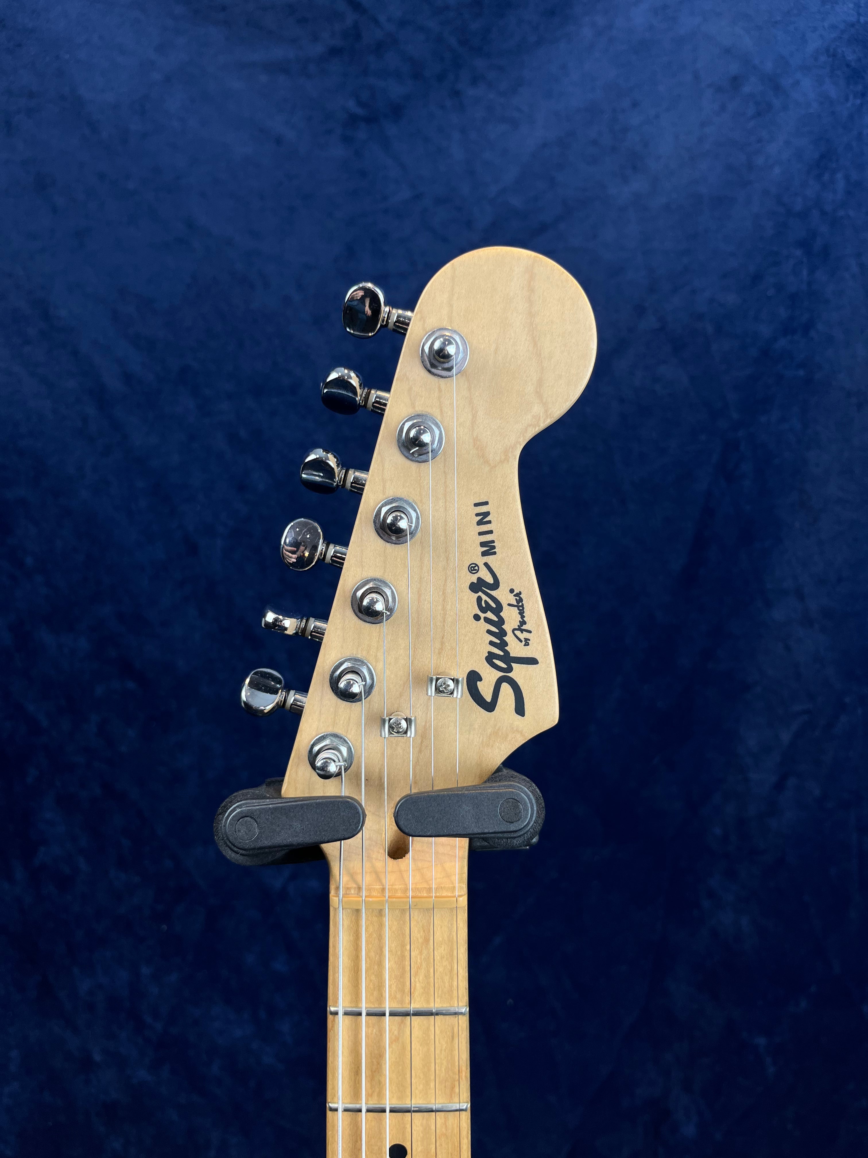 Light wood headstock of a Squier Mini electric guitar with six metal tuning pegs, black logo, and visible guitar strings against a dark blue background.