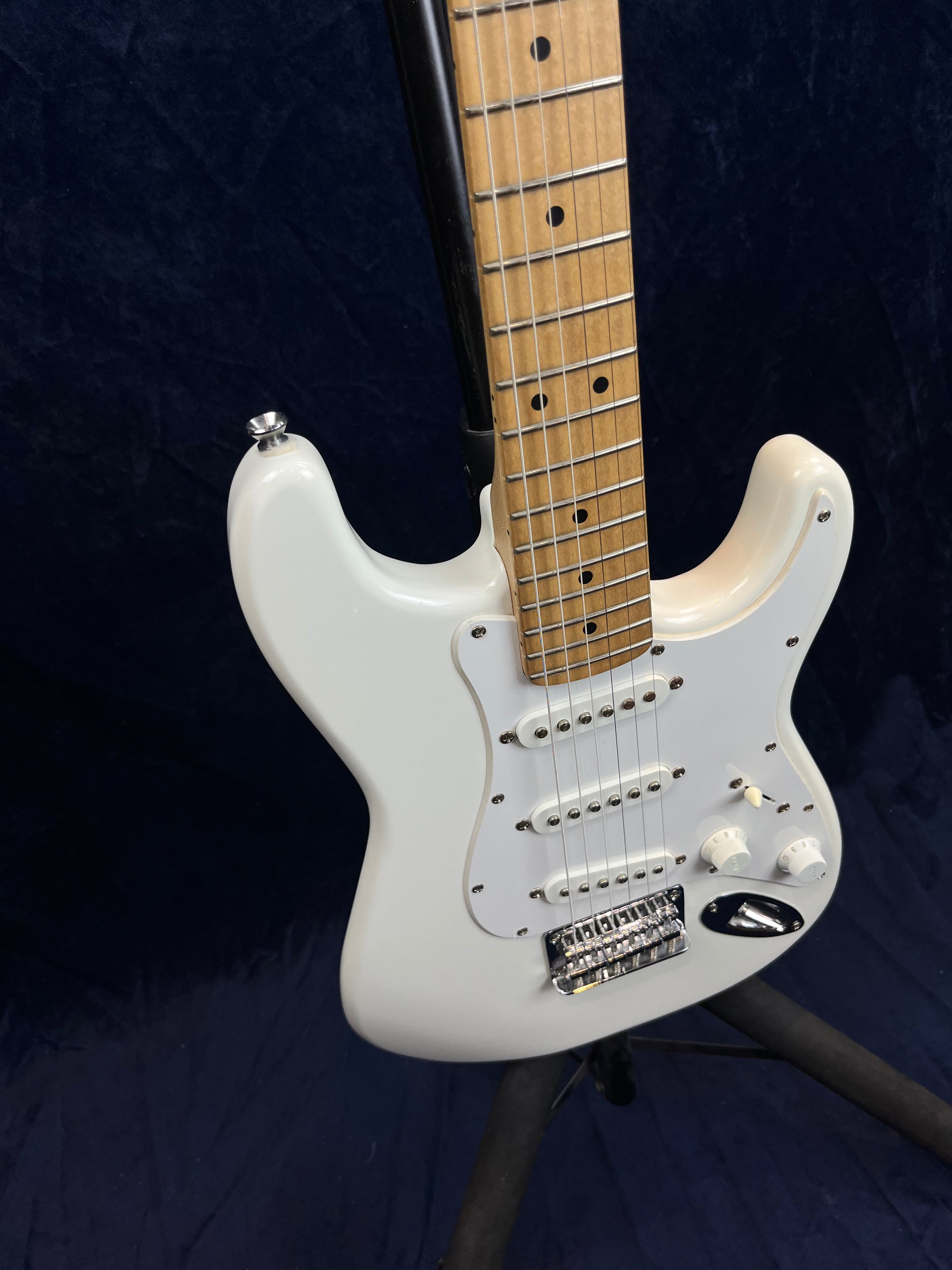 White electric guitar with three single-coil pickups, two control knobs, a pickup switch, and a light wood fretboard on a dark background.