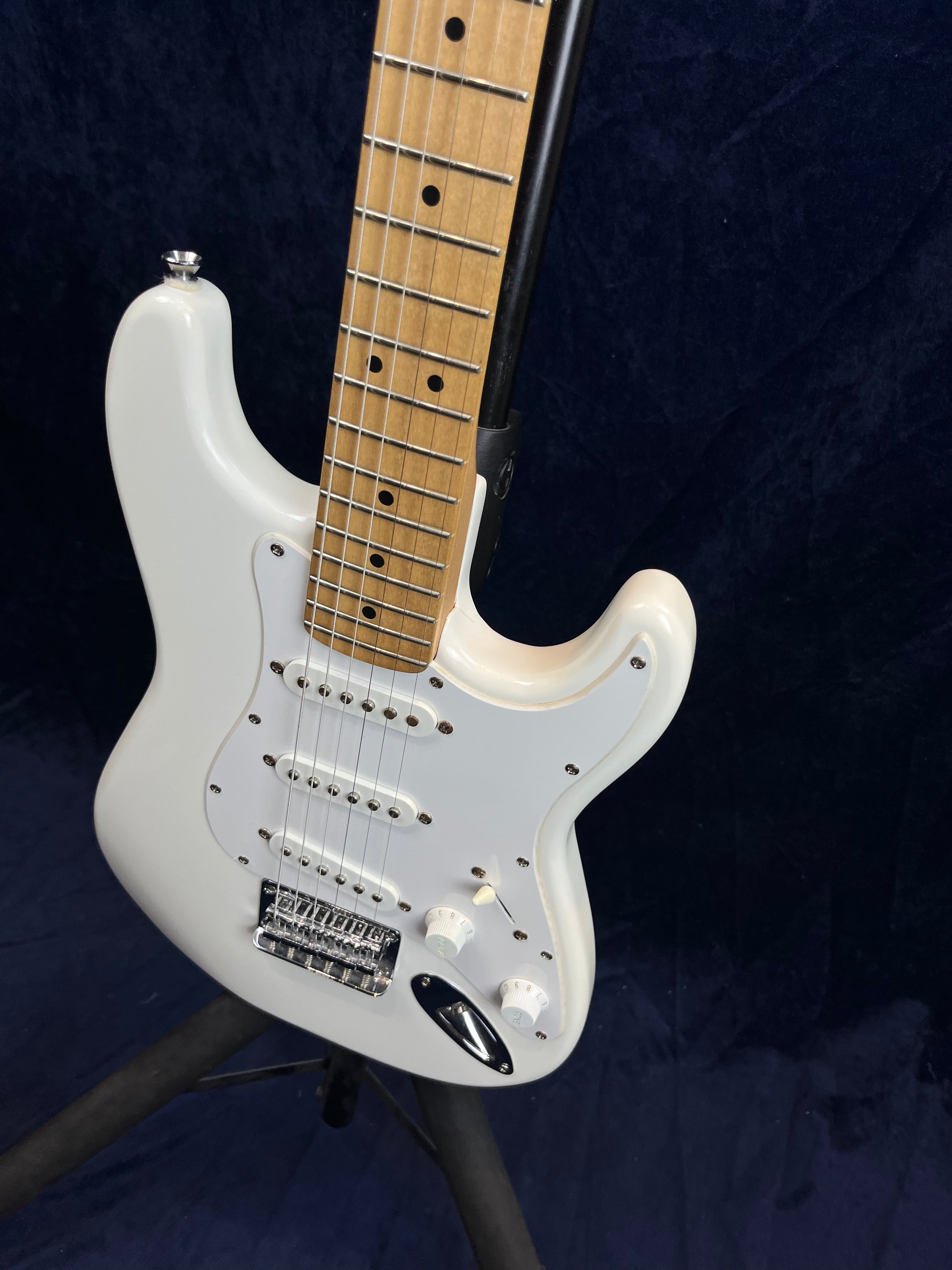 White electric guitar with a maple neck, three single-coil pickups, white pickguard, and three control knobs, displayed on a black stand.