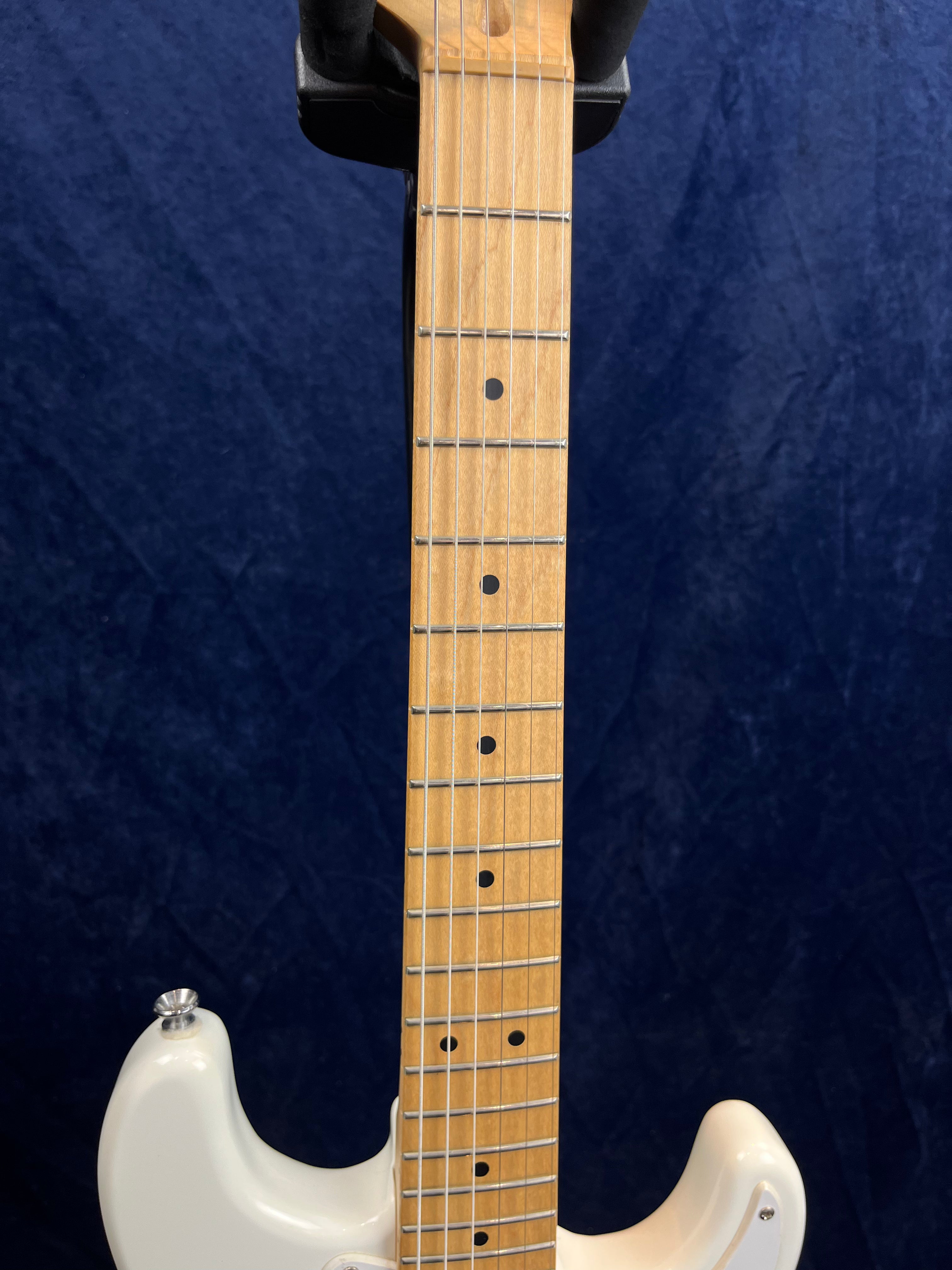 A light wood electric guitar neck with dot inlays and six strings is attached to a white guitar body, shown against a dark blue background.