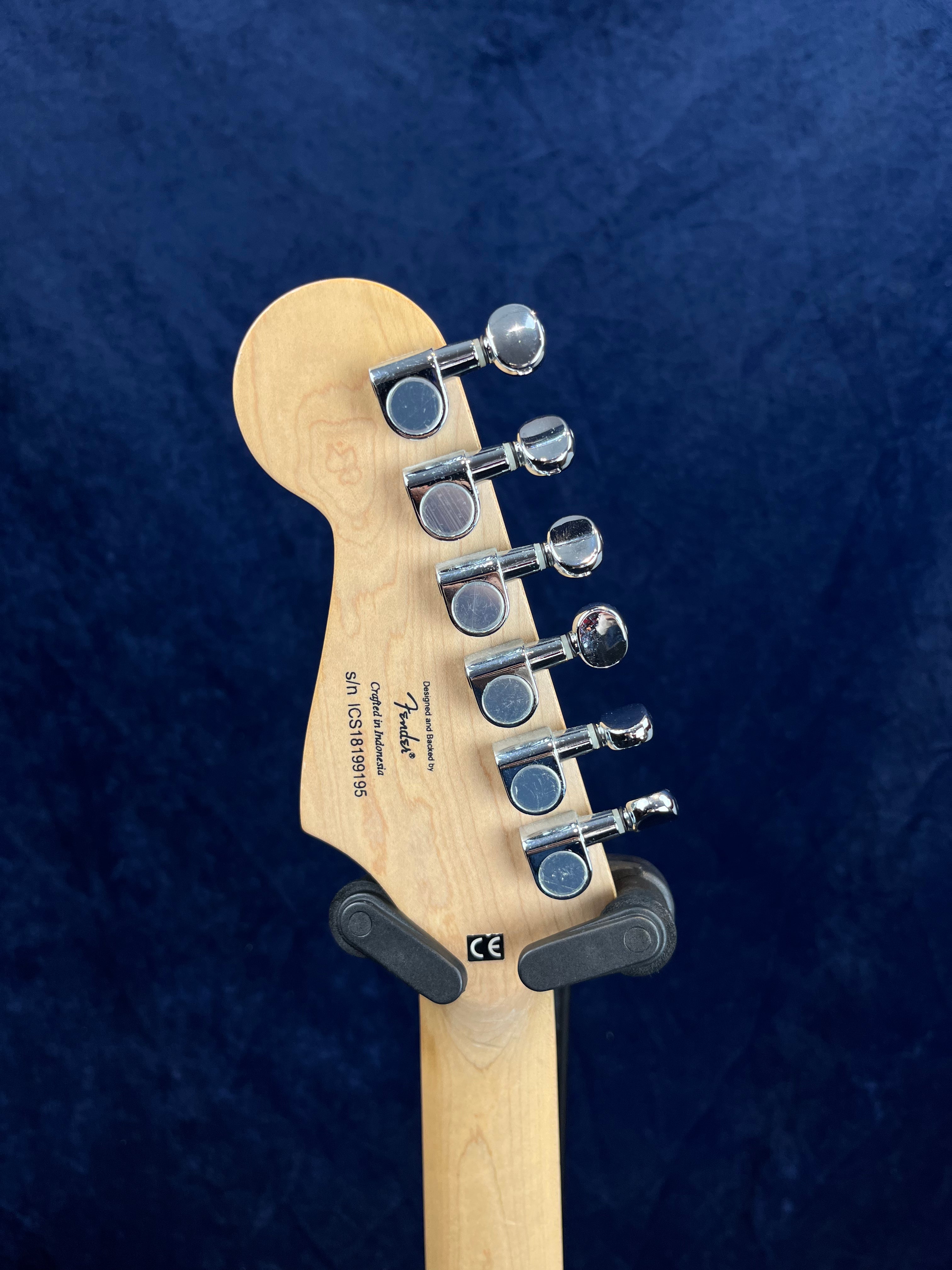 Back of a Fender electric guitar headstock with six chrome tuning pegs, serial number, and manufacturer's label visible against a dark blue background.