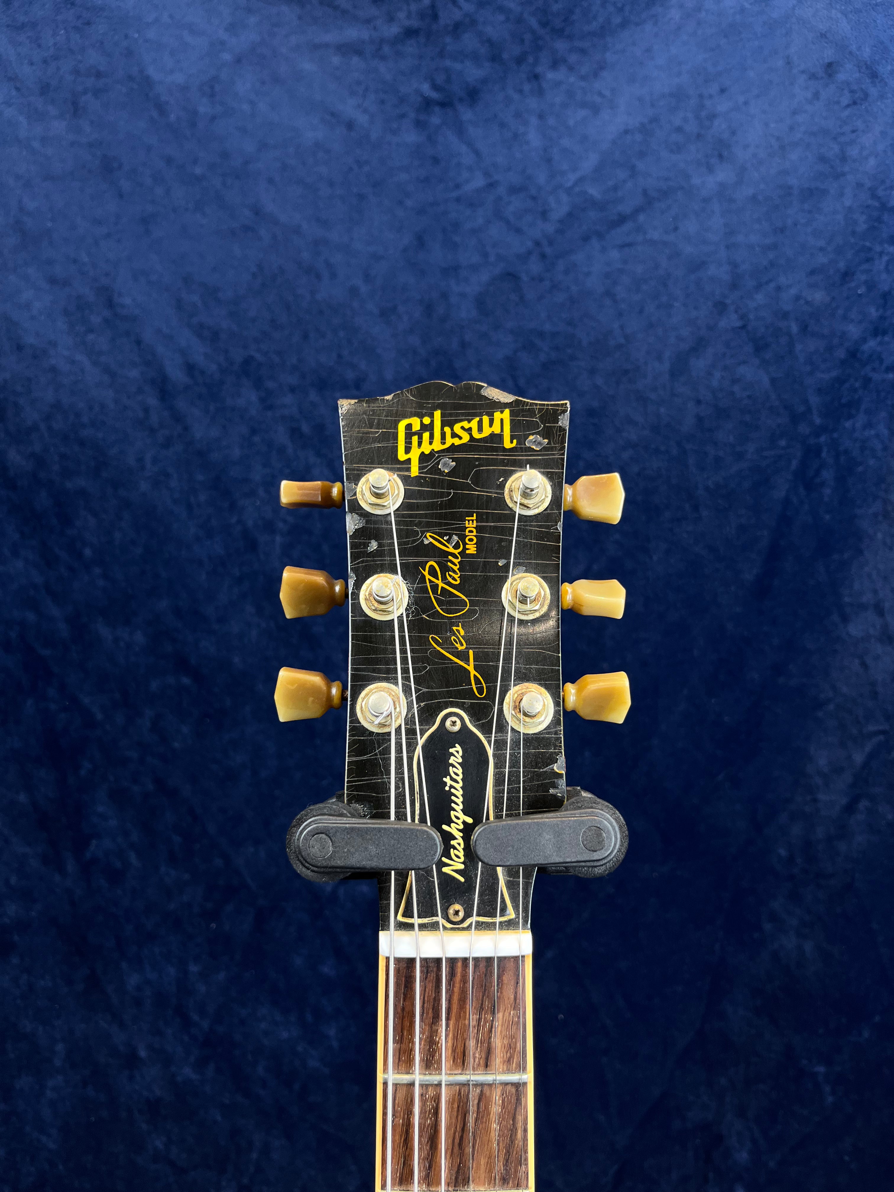 Close-up of a worn Gibson Les Paul electric guitar headstock with six tuning pegs, strings, and a capo attached, set against a blue background.