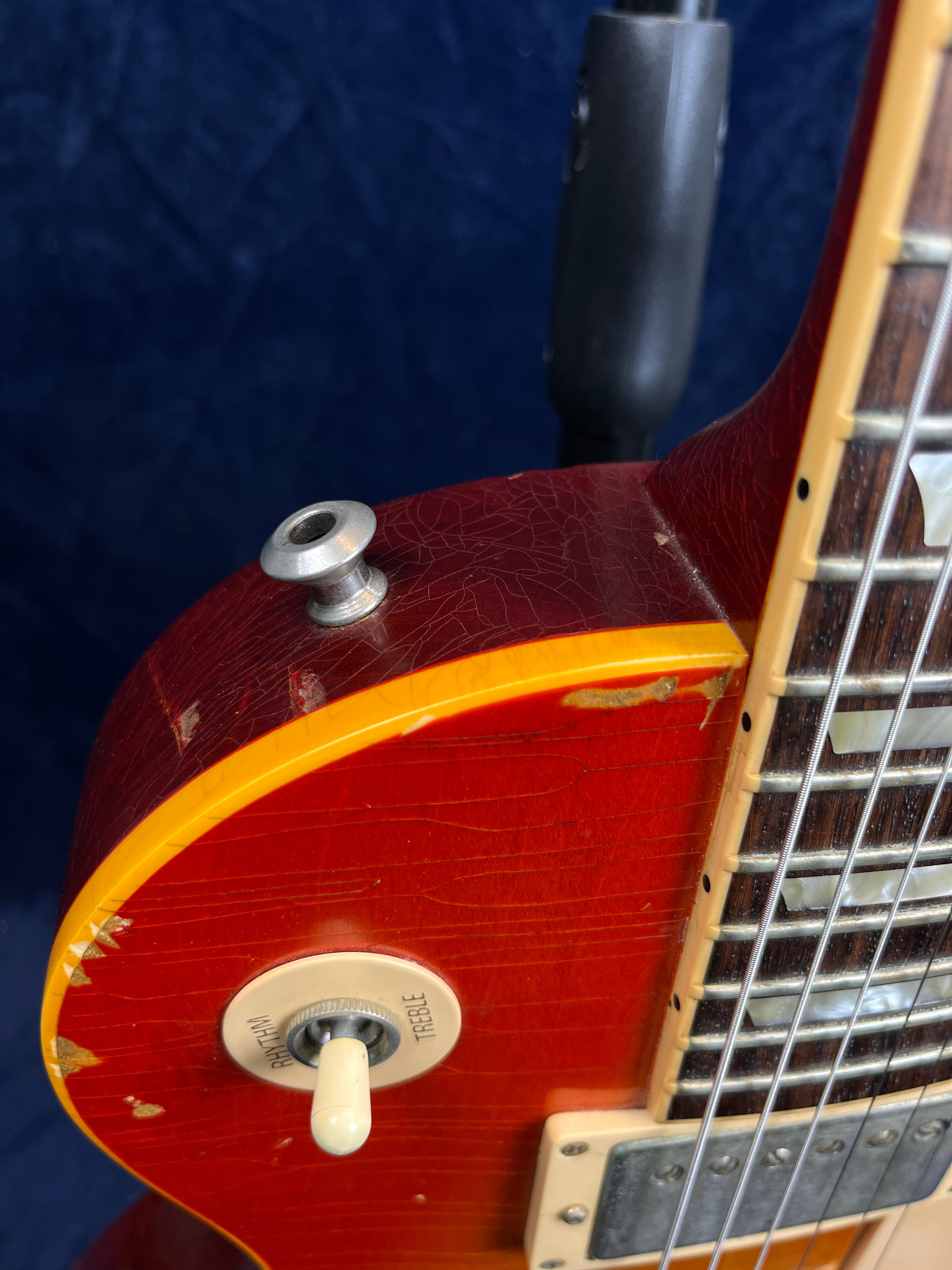 Electric guitar body close-up showing worn cherry-red finish, toggle switch labeled rhythm/treble, binding, and visible age cracks near the neck and edge.