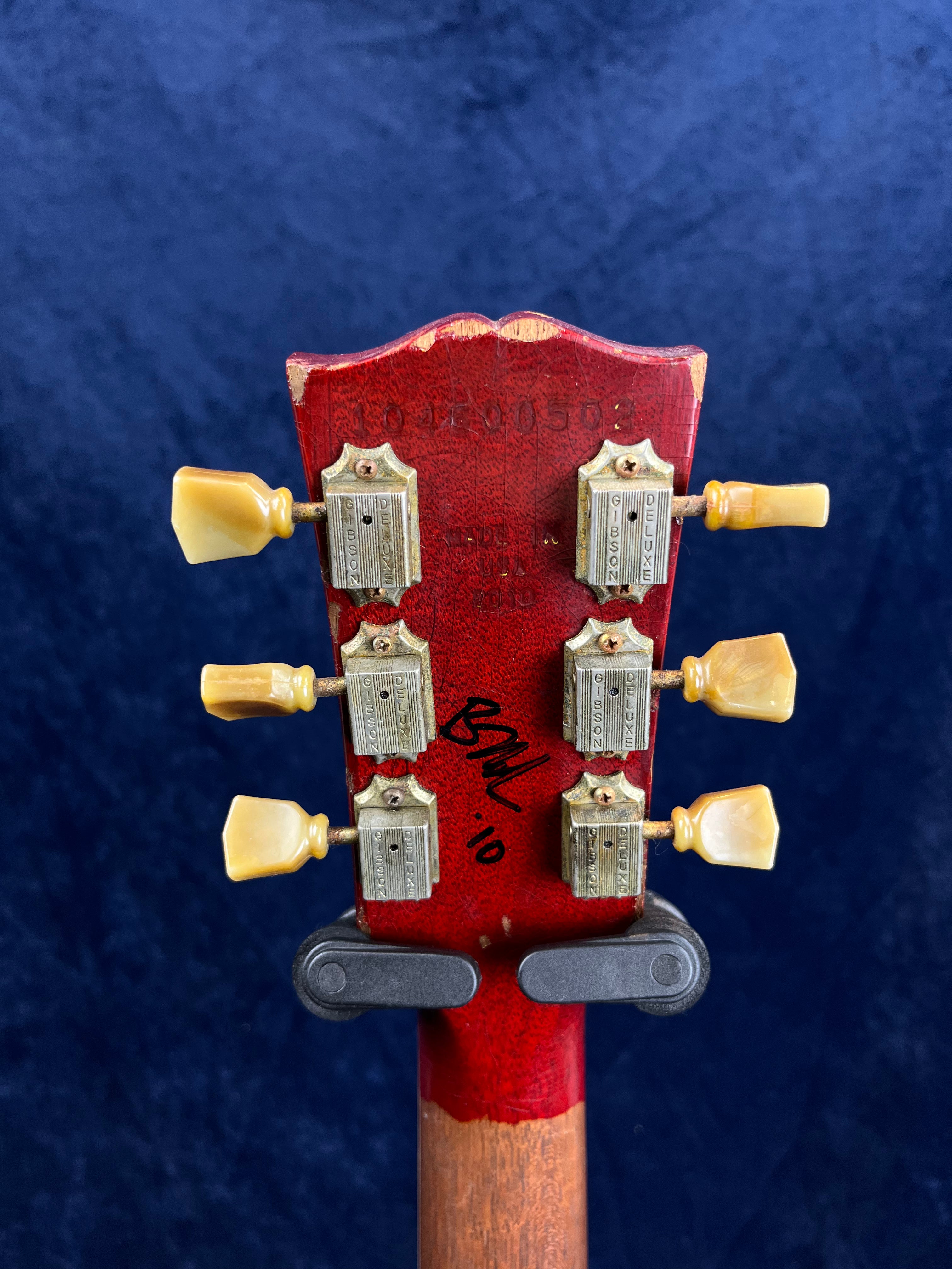 The back of a red electric guitar headstock shows six vintage-style tuners, visible serial numbers, a signature in black marker, and some chipped paint.
