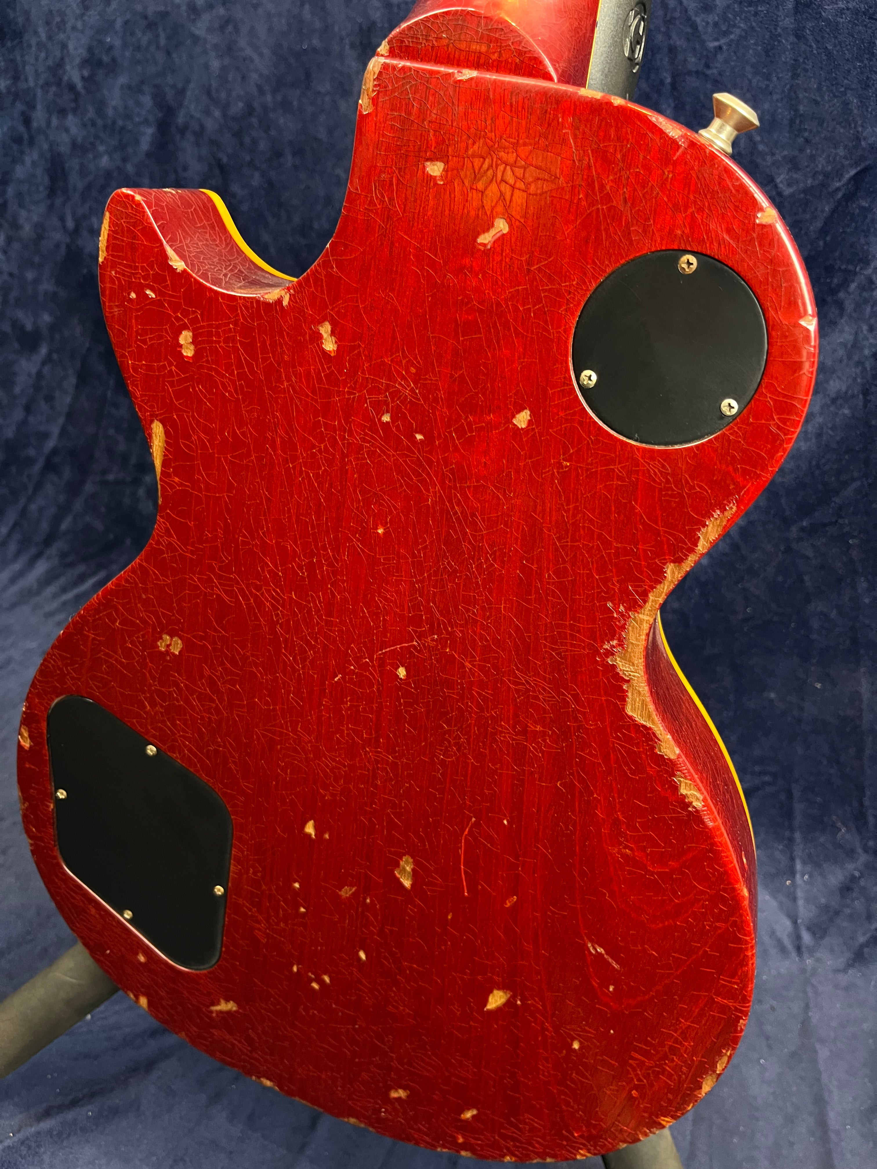 Electric guitar with a red cracked and worn finish, exposing wood in spots, two black back plates, and heavy relic wear around the edges.