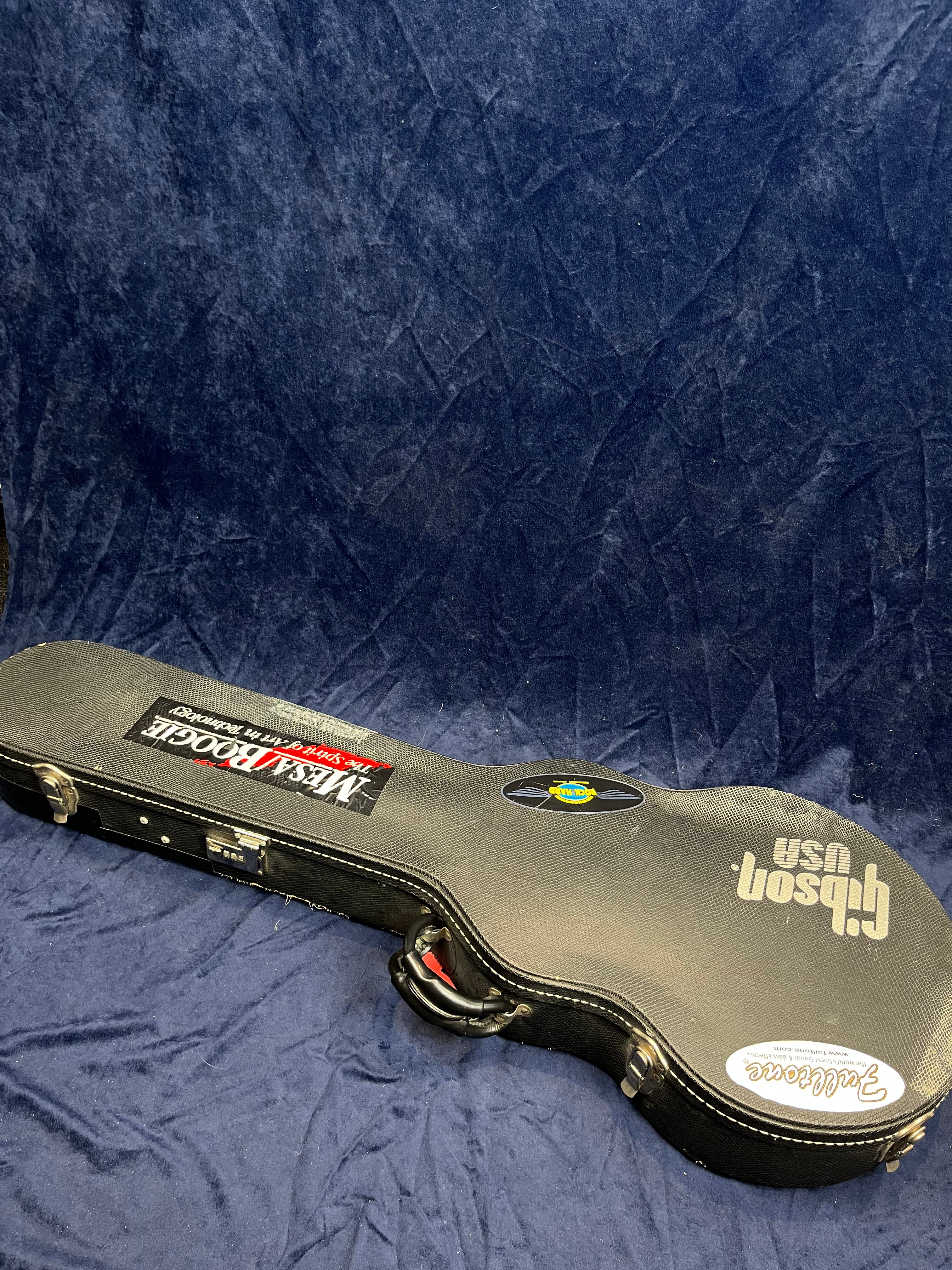 A hard shell electric guitar case with Gibson USA, Mesa/Boogie, and other stickers, resting on a dark blue textured surface.