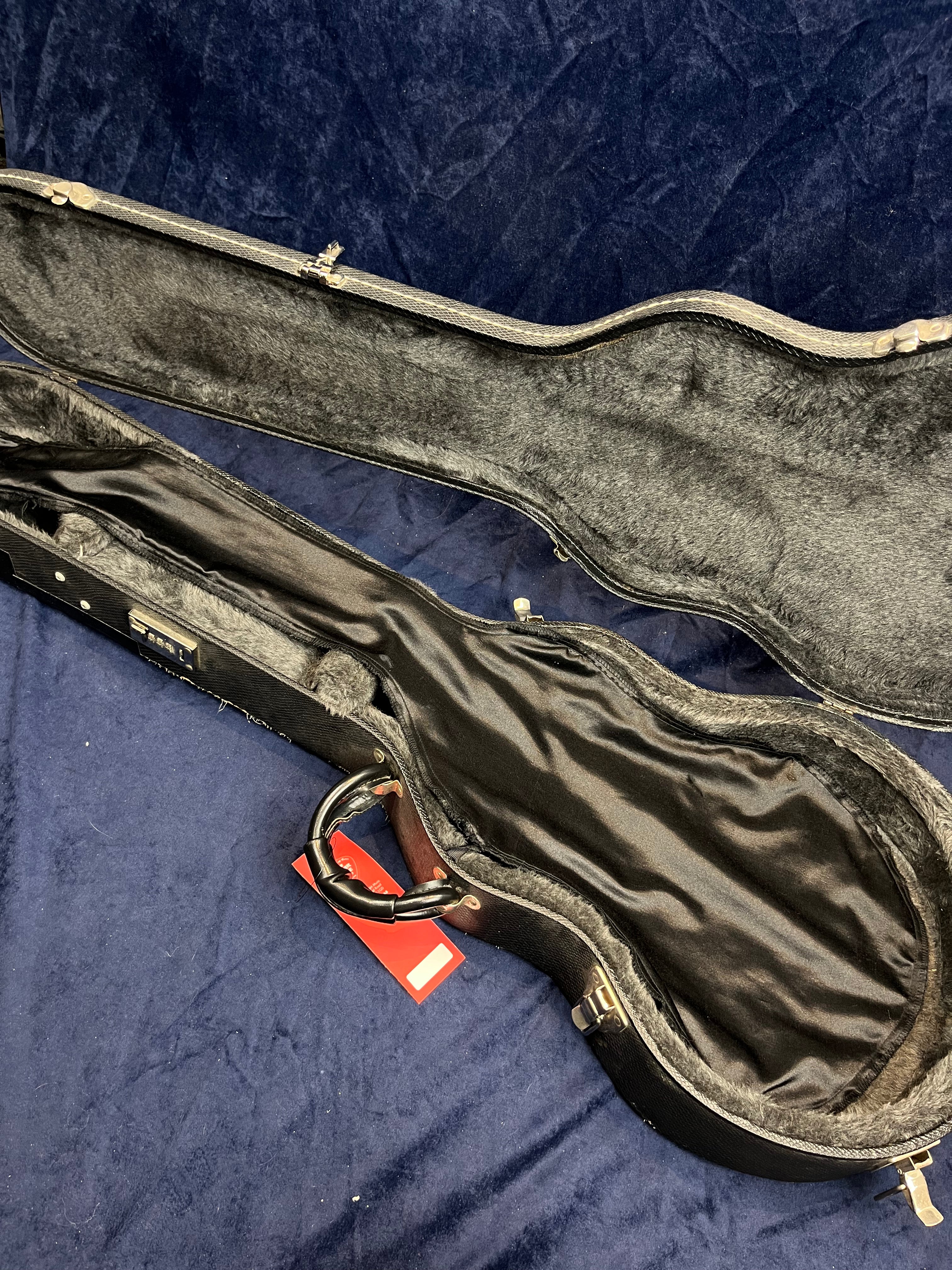 Open empty electric guitar case with a black plush interior and a black handle, sitting on a dark blue surface. A red tag is attached to the handle.