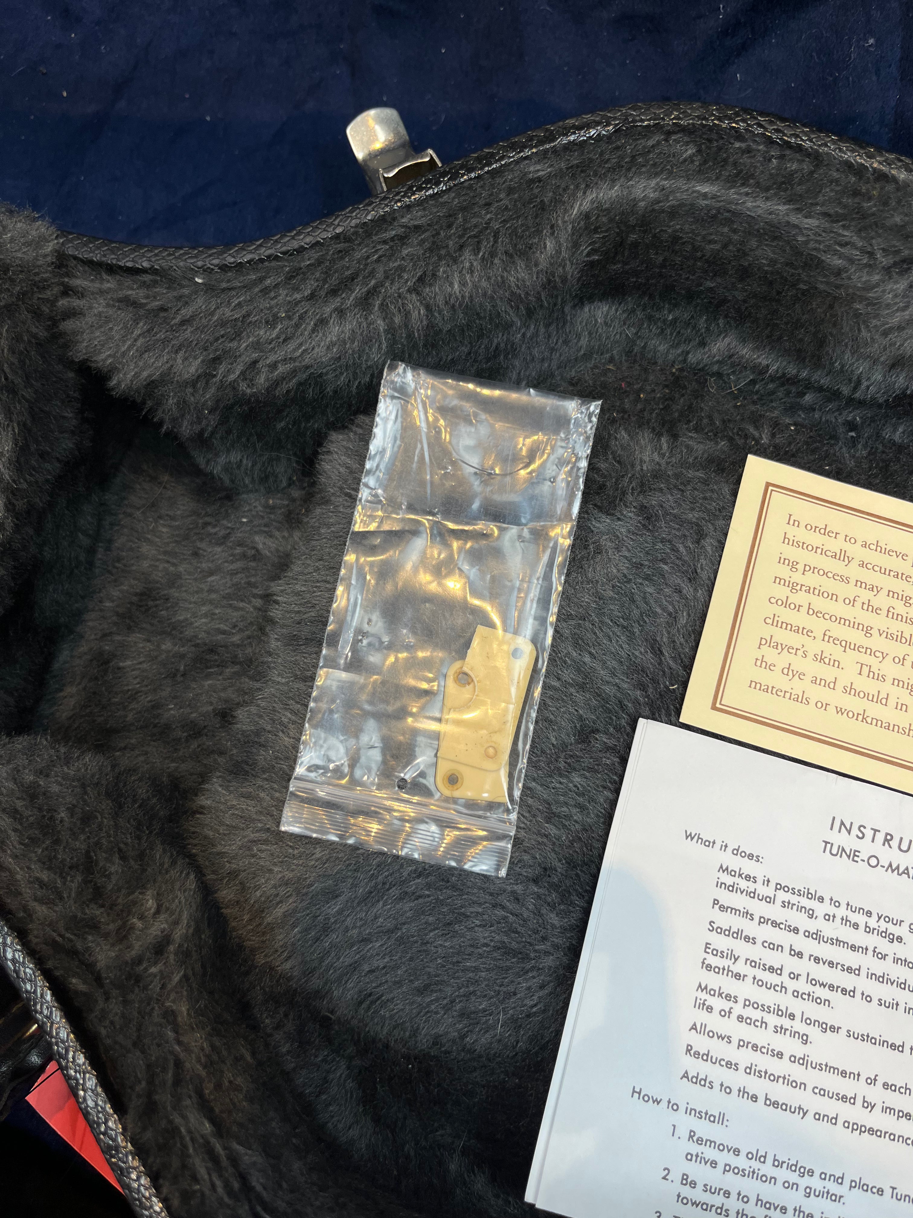 A sealed plastic bag containing a small cream-colored guitar part rests in a plush black electric guitar case beside folded instruction sheets.