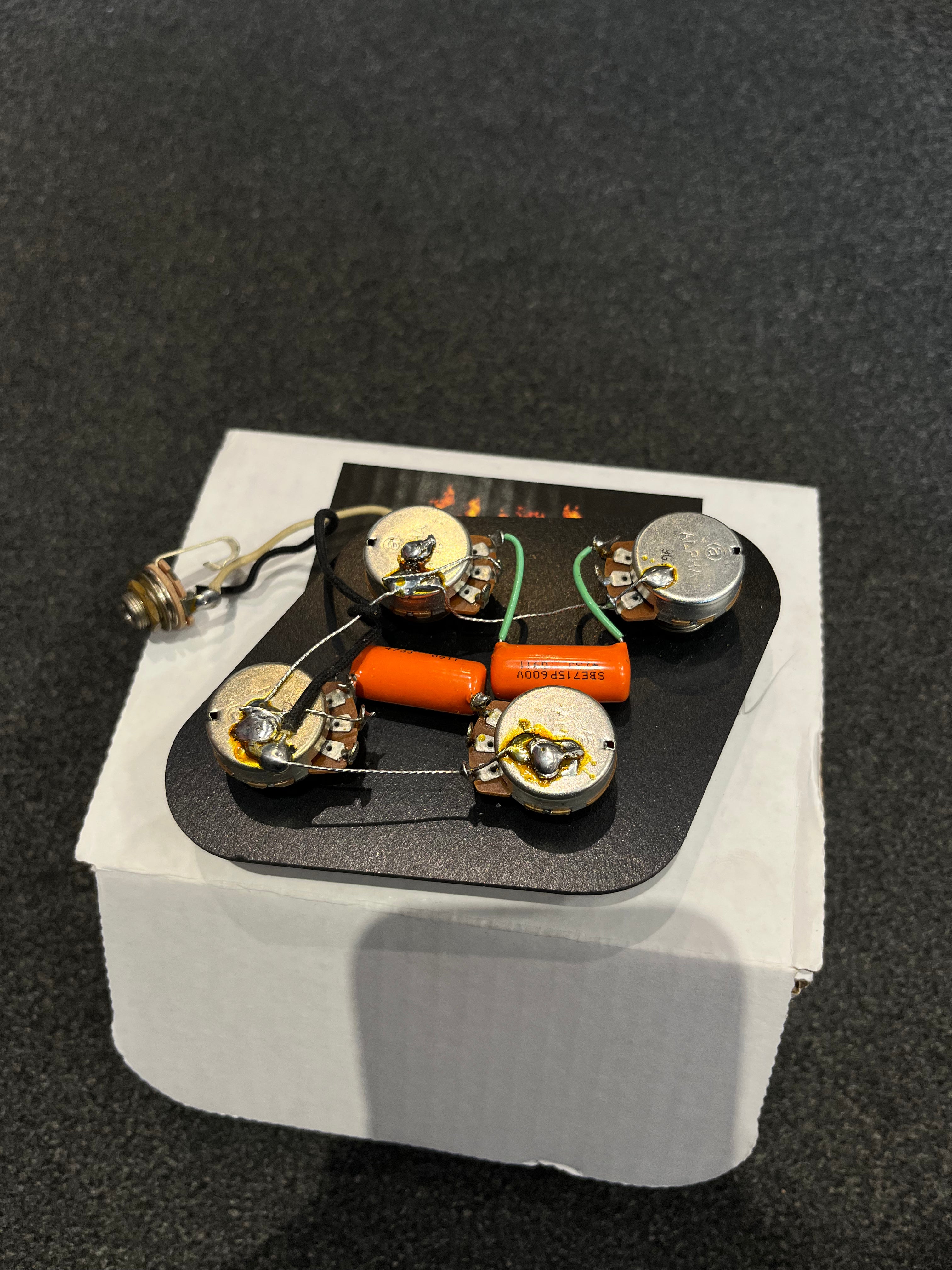 Electric guitar wiring harness with four potentiometers, an orange capacitor, and attached wires, resting on a small white box.