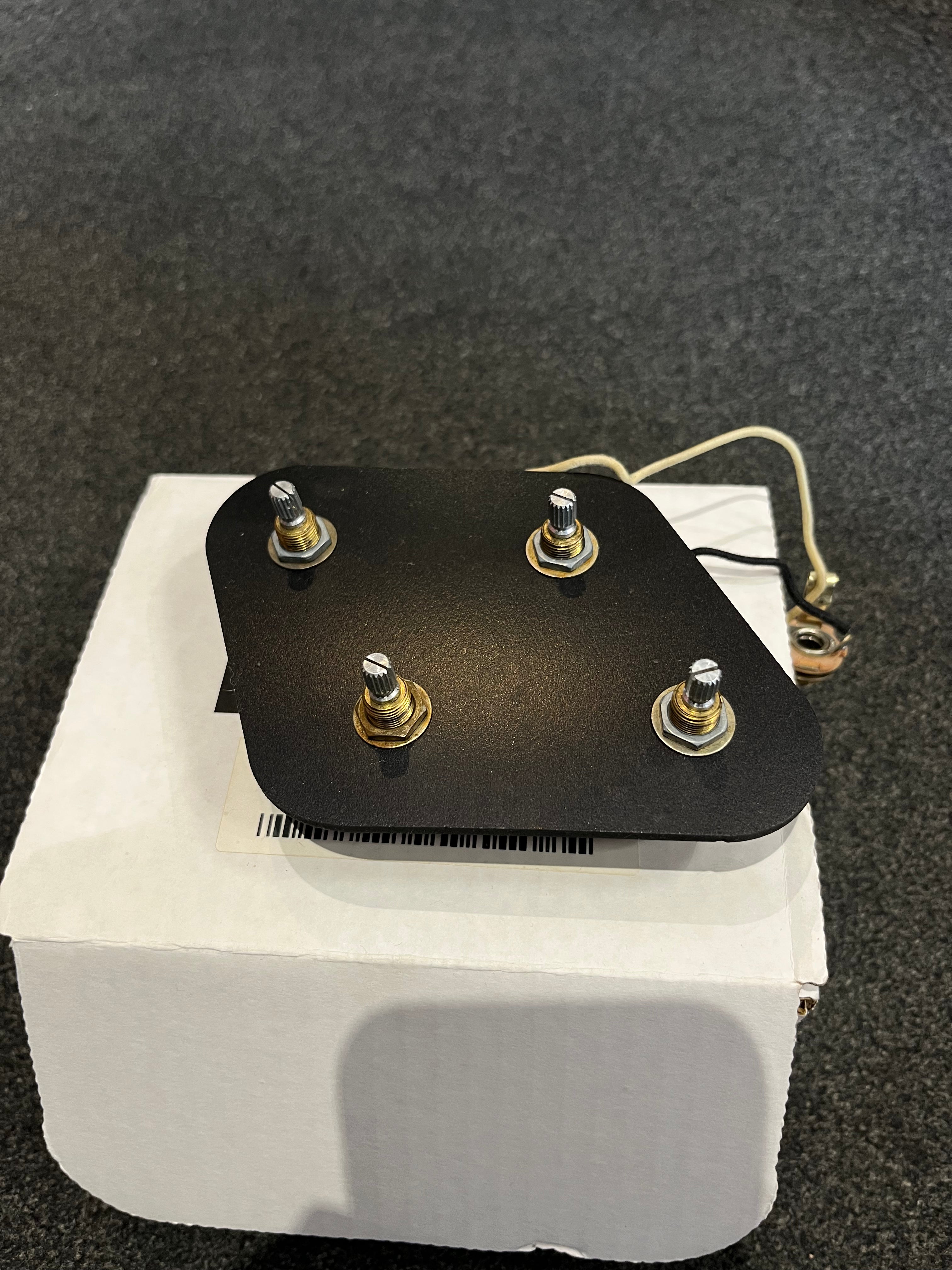 Electric guitar control plate with four metal knob posts and attached wires rests on a white box, placed on a dark textured surface.