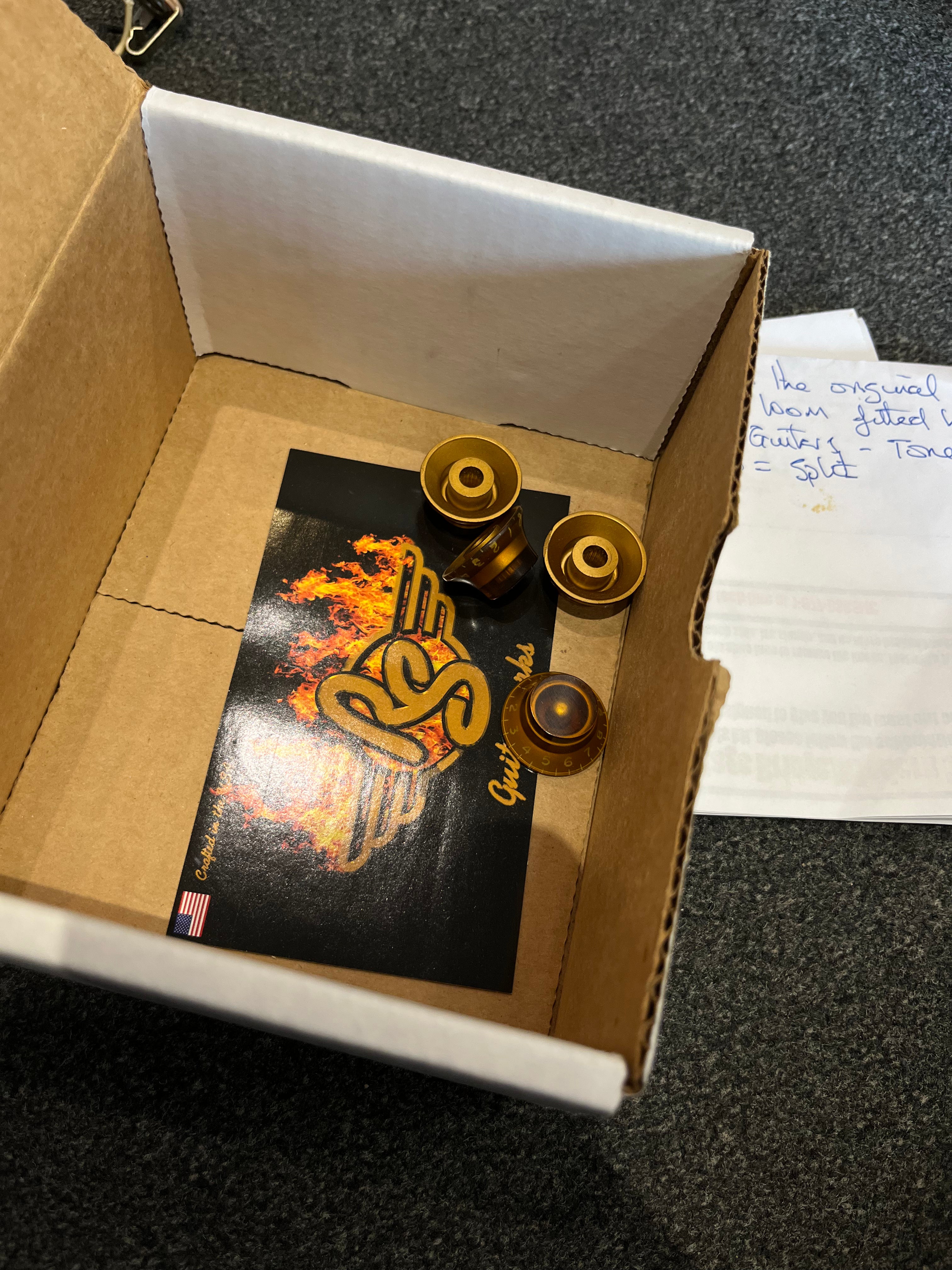 Three amber-colored electric guitar control knobs and a card with a flaming logo are inside an open cardboard box on a dark countertop.