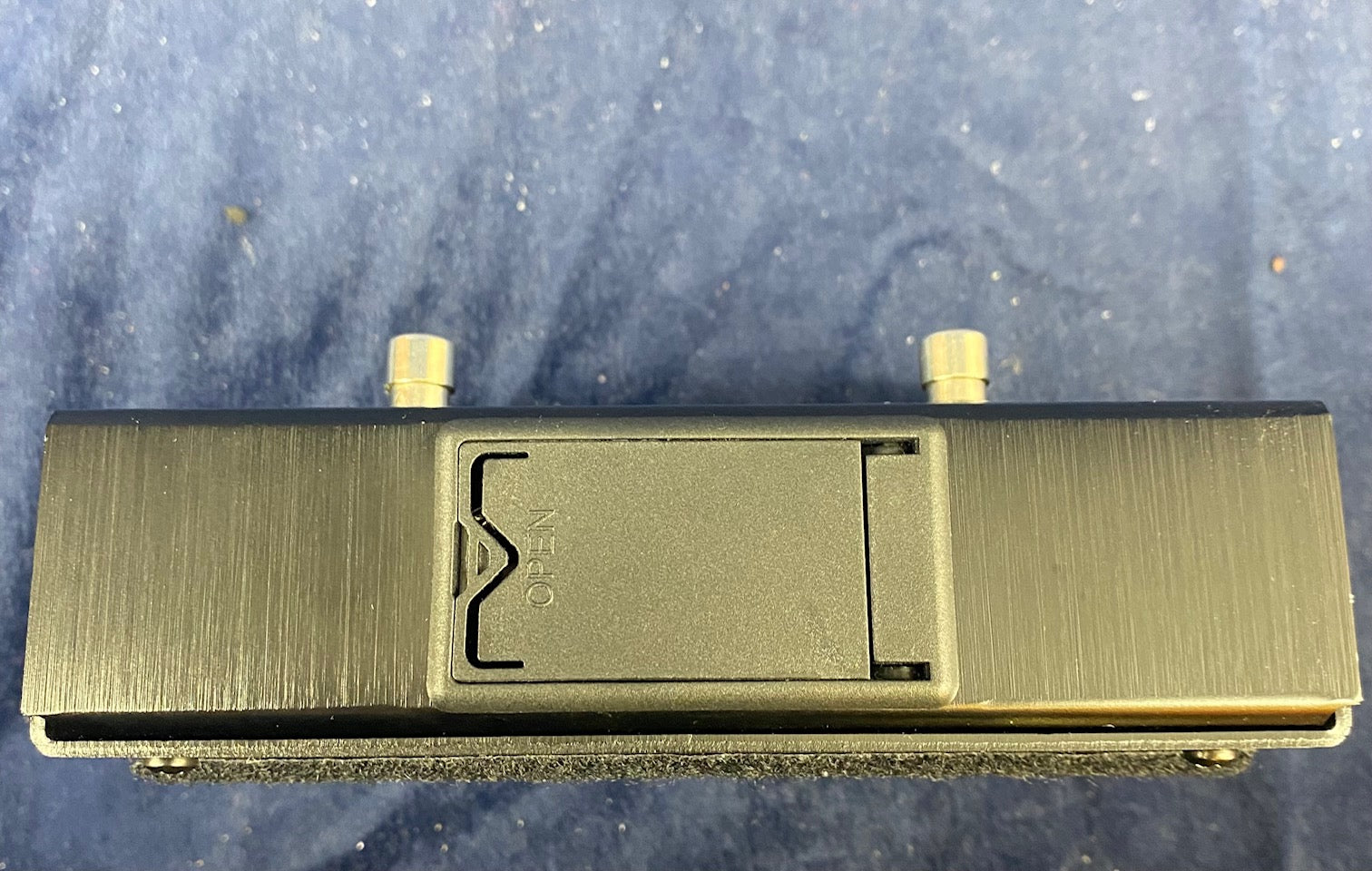 Rectangular FX pedal with brushed black surface, a central battery compartment labeled "open," two metal footswitches on each end, on a blue background.