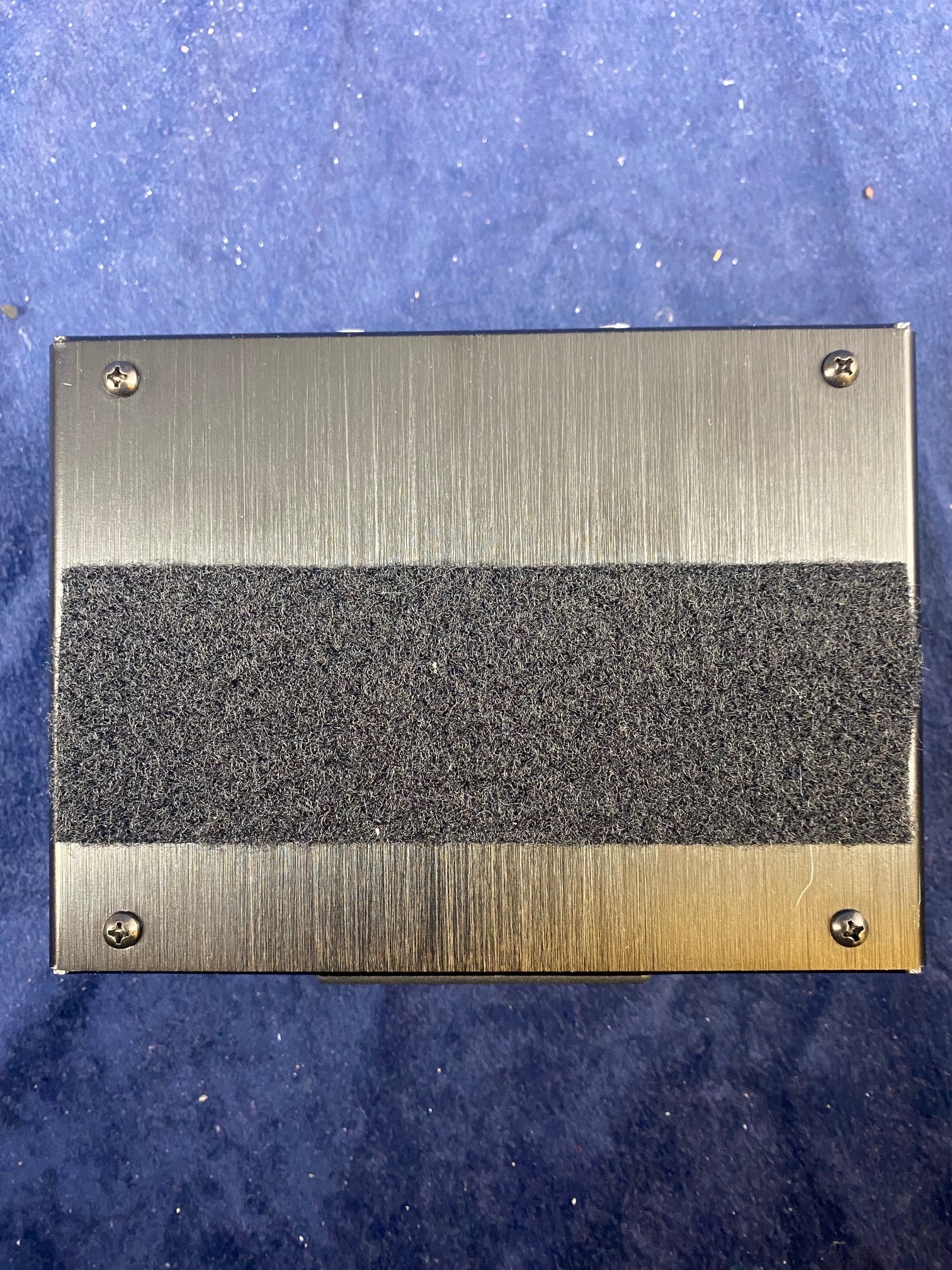 The bottom of an FX pedal with a brushed metal finish, four corner screws, and a wide strip of black hook-and-loop fastener across the center.