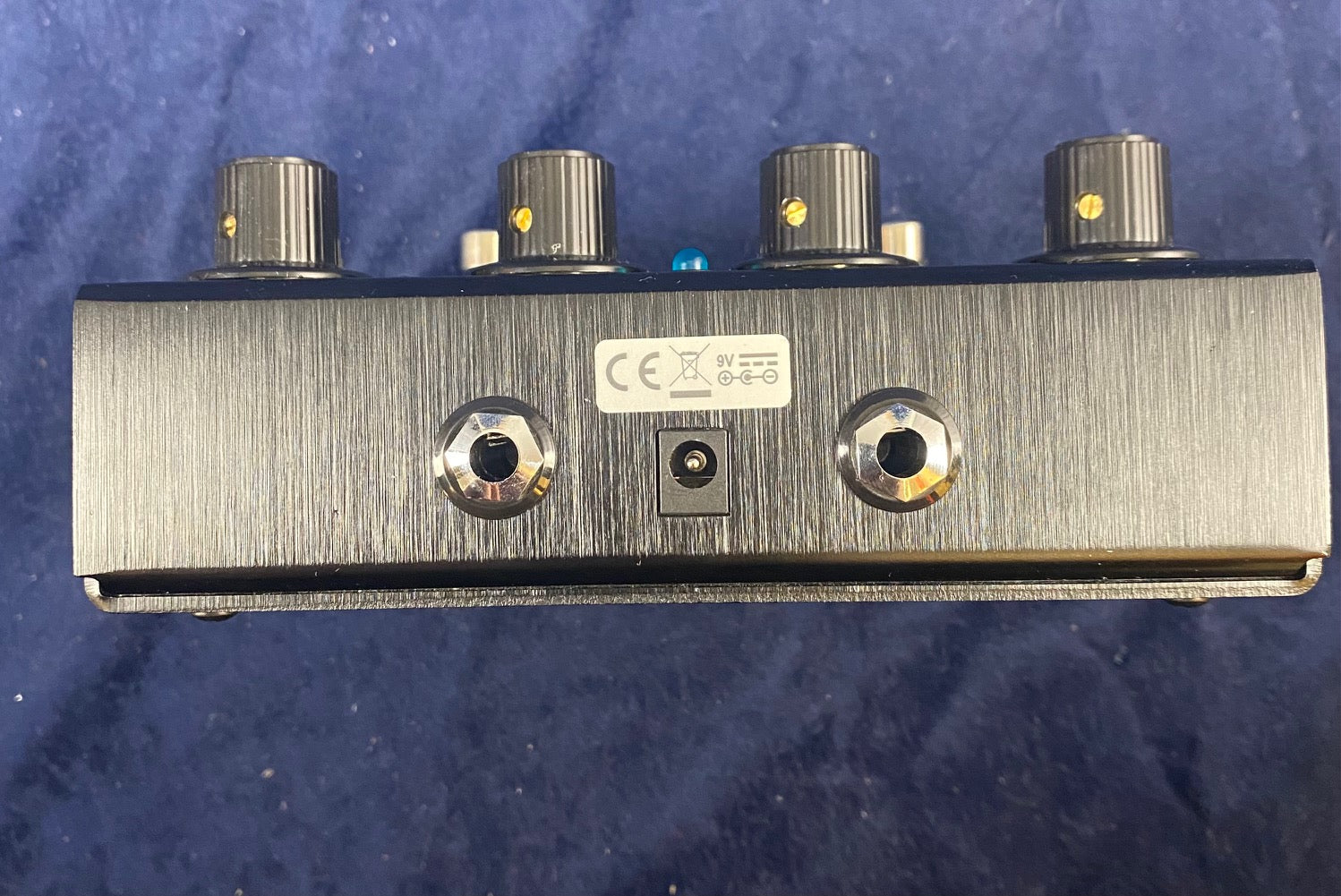 FX pedal showing three audio jacks and a power jack on a brushed metal panel, with four black control knobs on top.