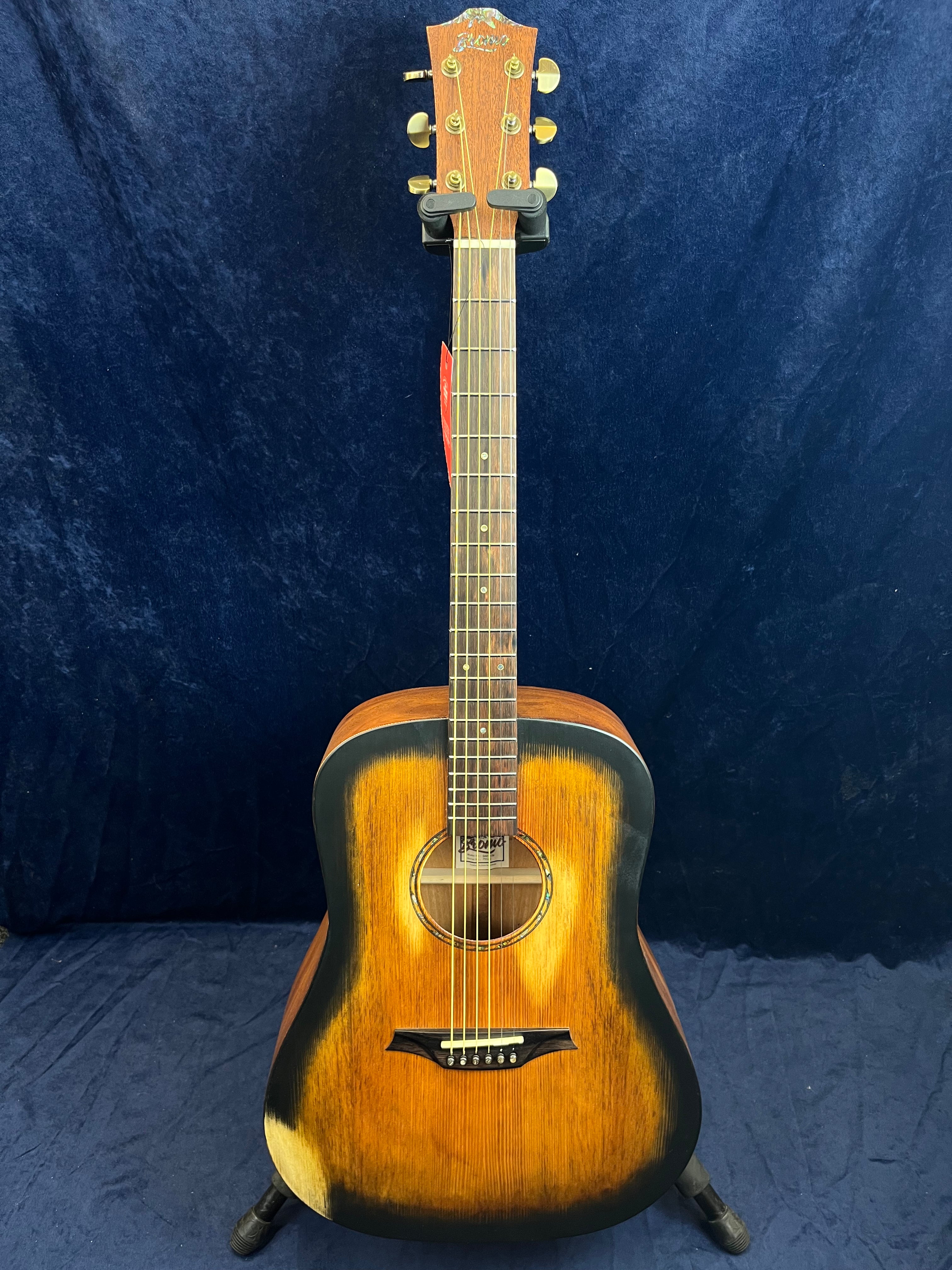 Sunburst acoustic guitar with a dark outer edge and lighter brown center, six strings, gold tuning pegs, and a unique shaped black bridge.