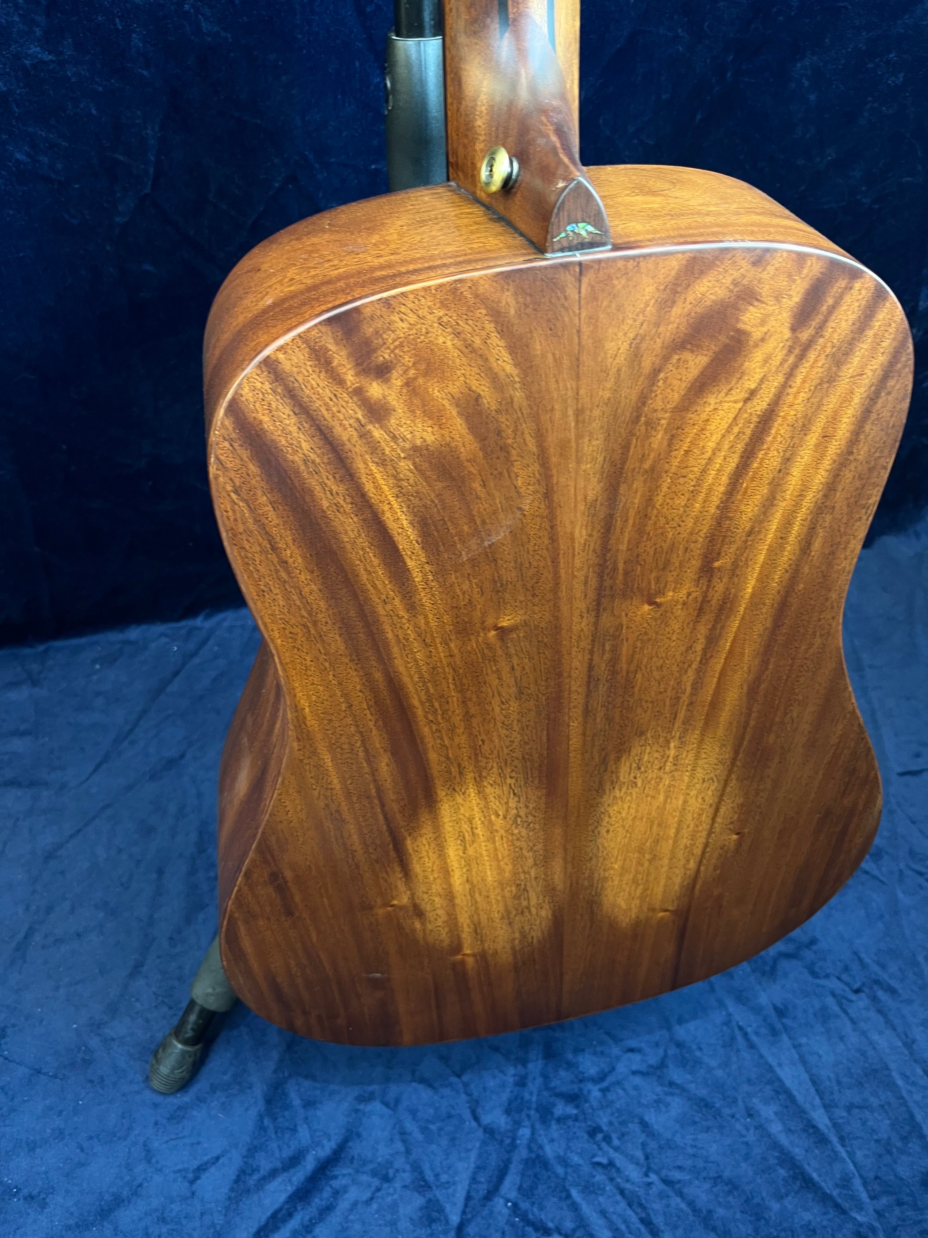 Back view of an acoustic guitar made from rich brown wood with a visible grain pattern, resting on a stand against a dark blue background.