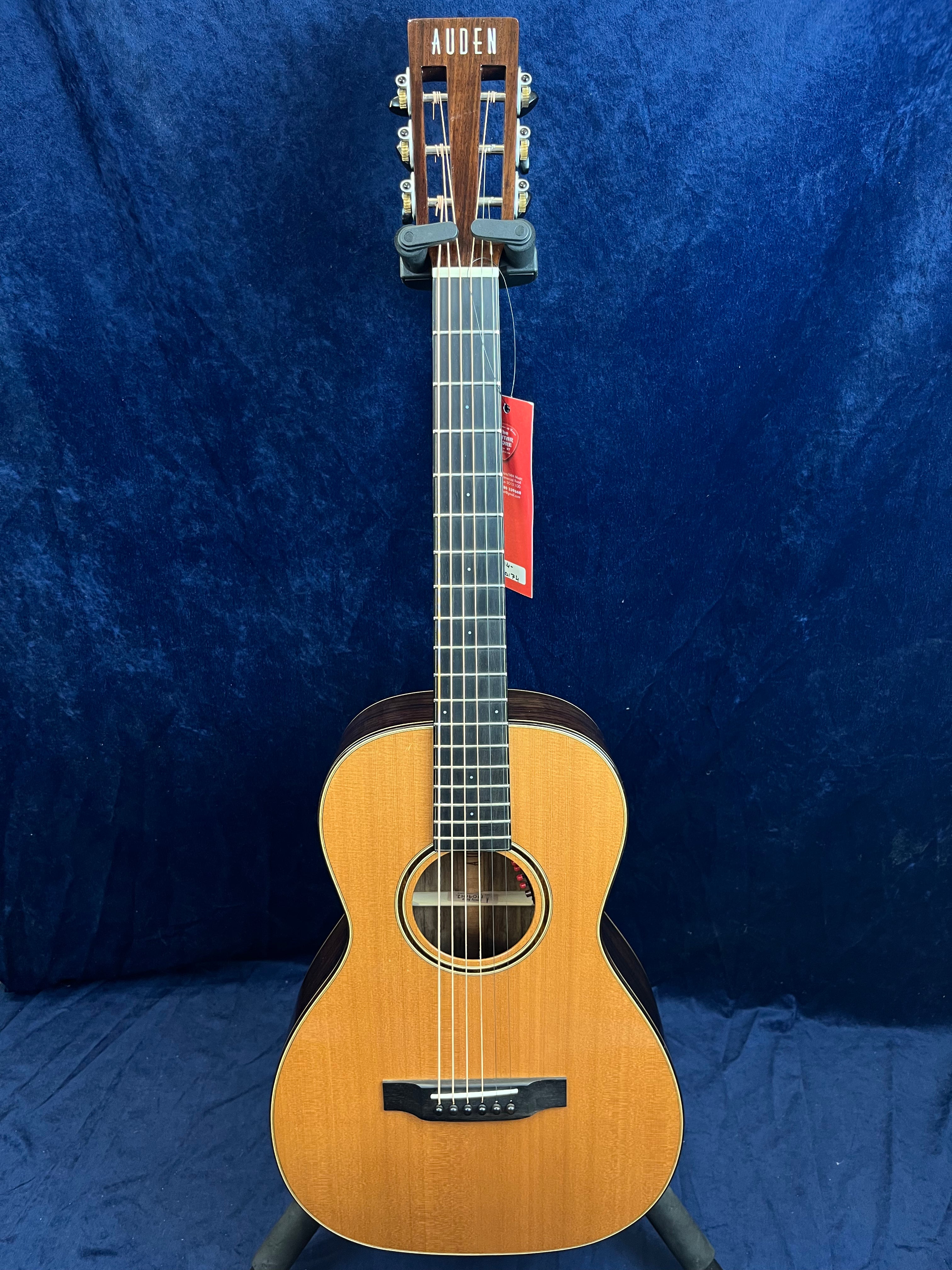 Auden acoustic guitar with a natural wood finish, dark fretboard, and slotted headstock, displayed upright against a dark blue background.