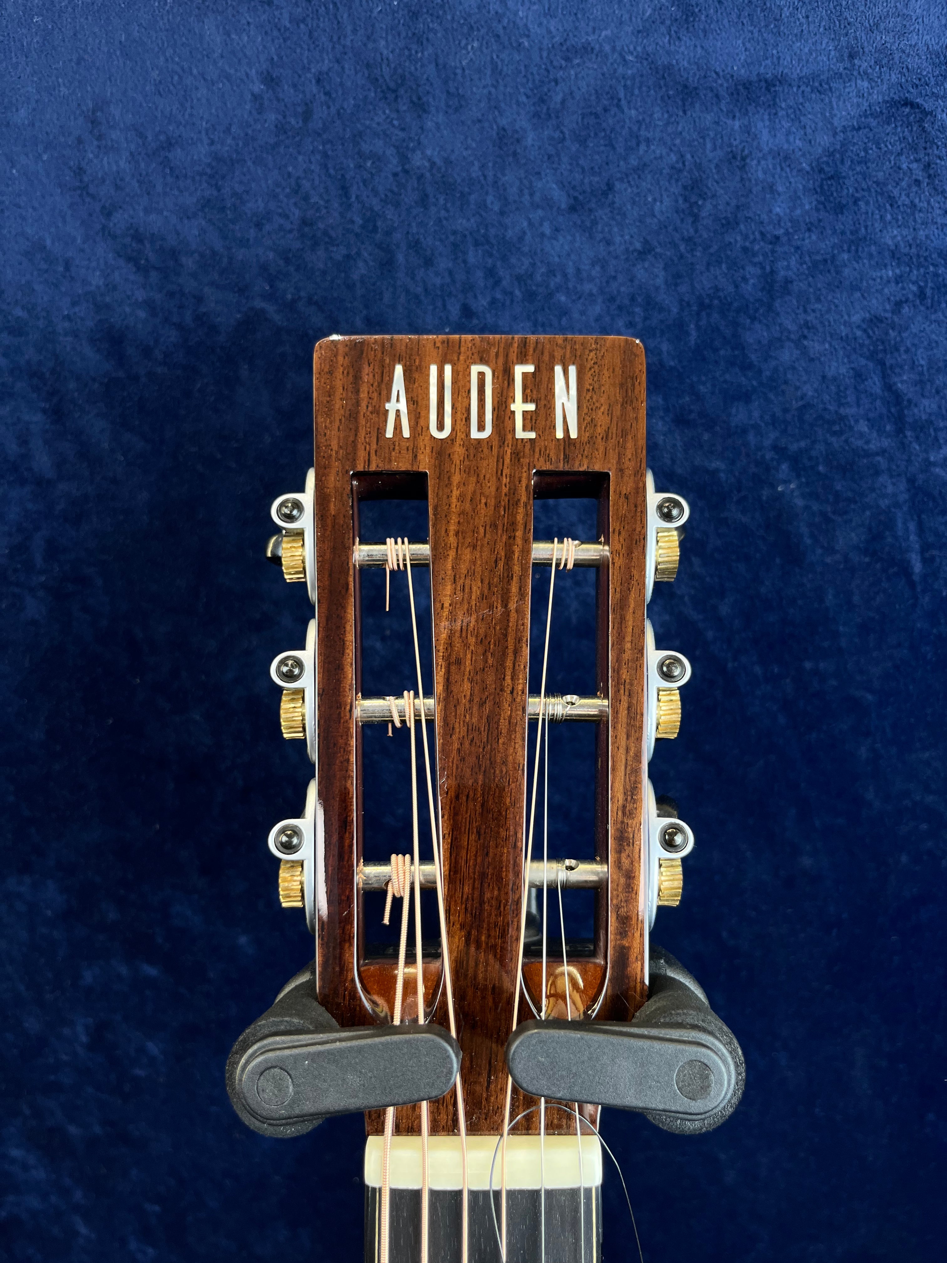 Auden acoustic guitar headstock with gold tuning pegs, visible string windings, and a dark wood finish against a dark blue background.