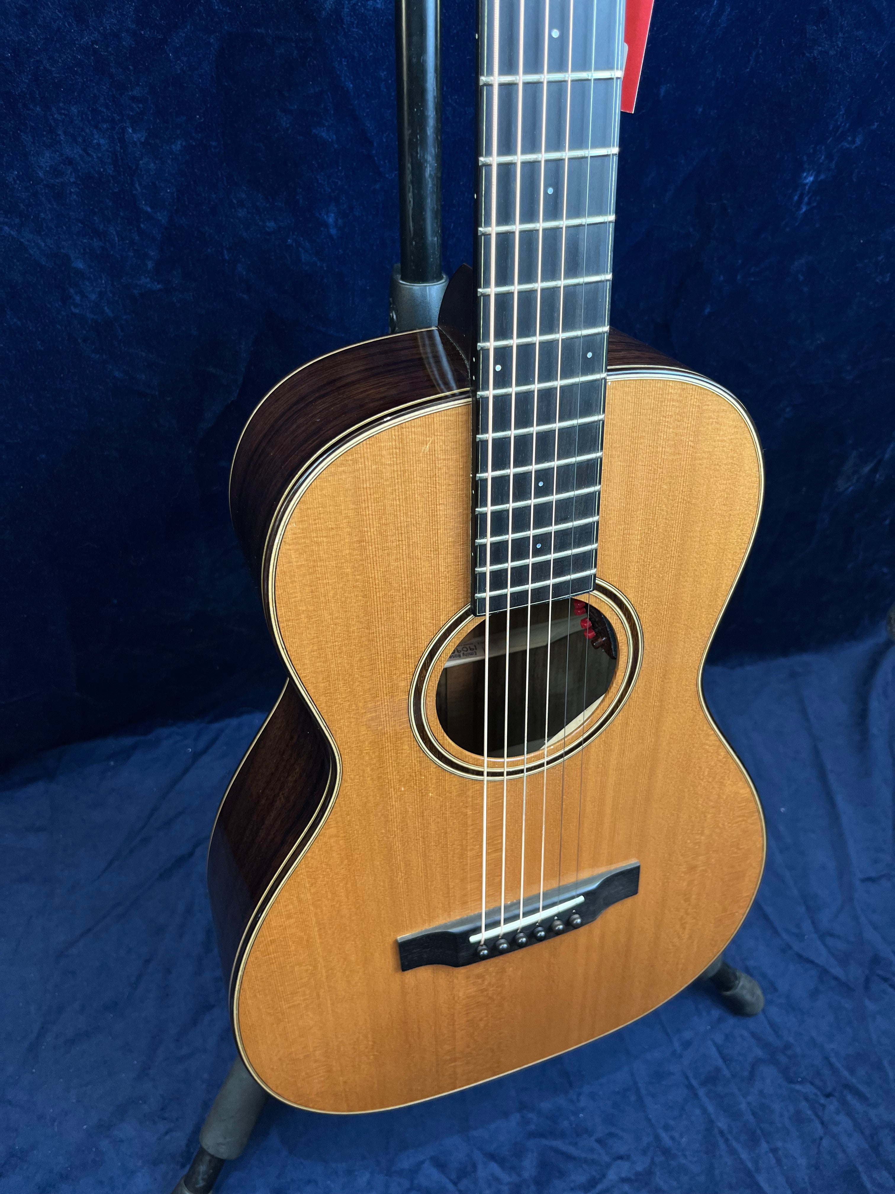 Small-bodied acoustic guitar with a natural wood top, dark sides, six strings, and a simple round soundhole, shown upright on a stand.