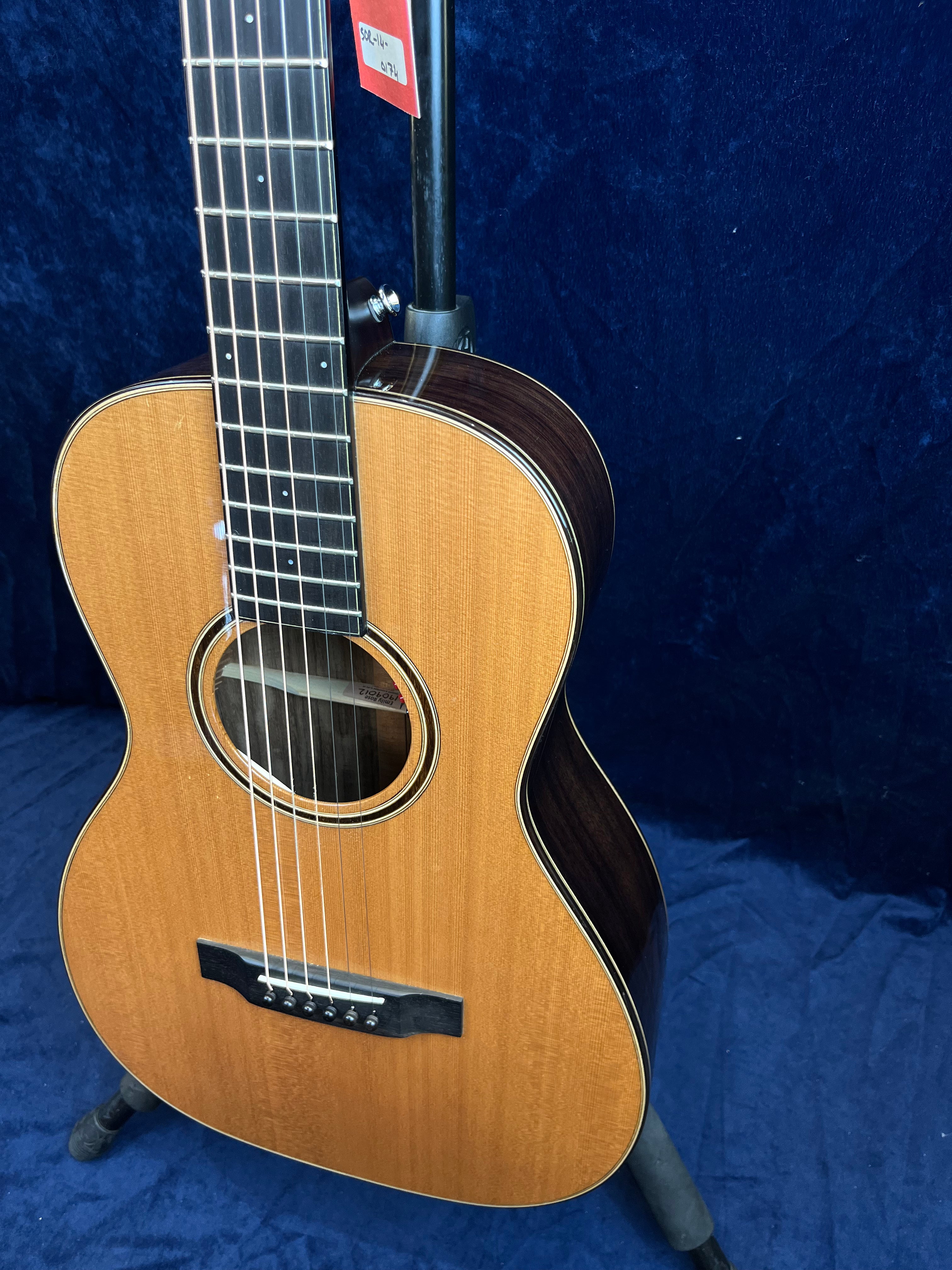 Small-bodied acoustic guitar with a natural wood top, dark wood sides, six strings, and dot inlays on the fretboard, displayed on a stand.