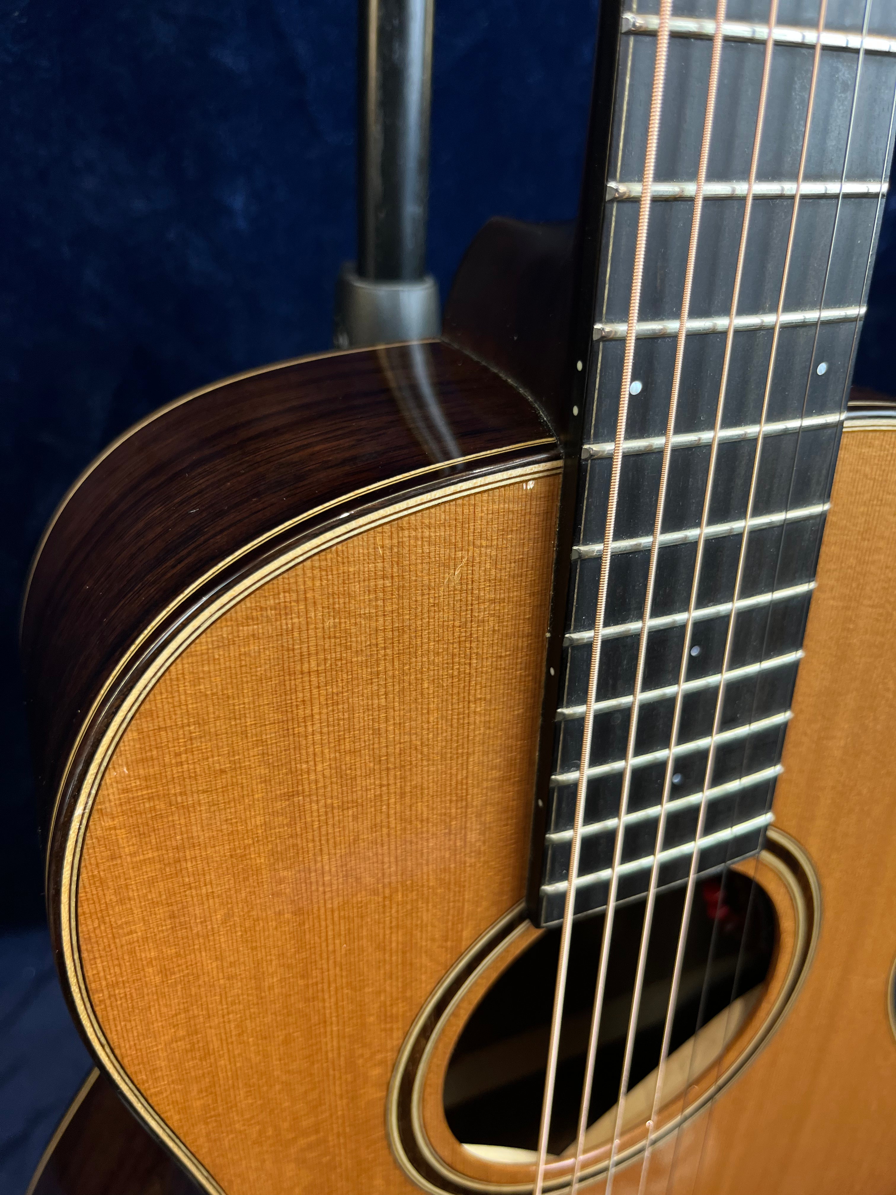 Close-up of an acoustic guitar’s body and strings, showing a wooden top, dark wood sides, and part of the fingerboard with metal frets and dot inlays.