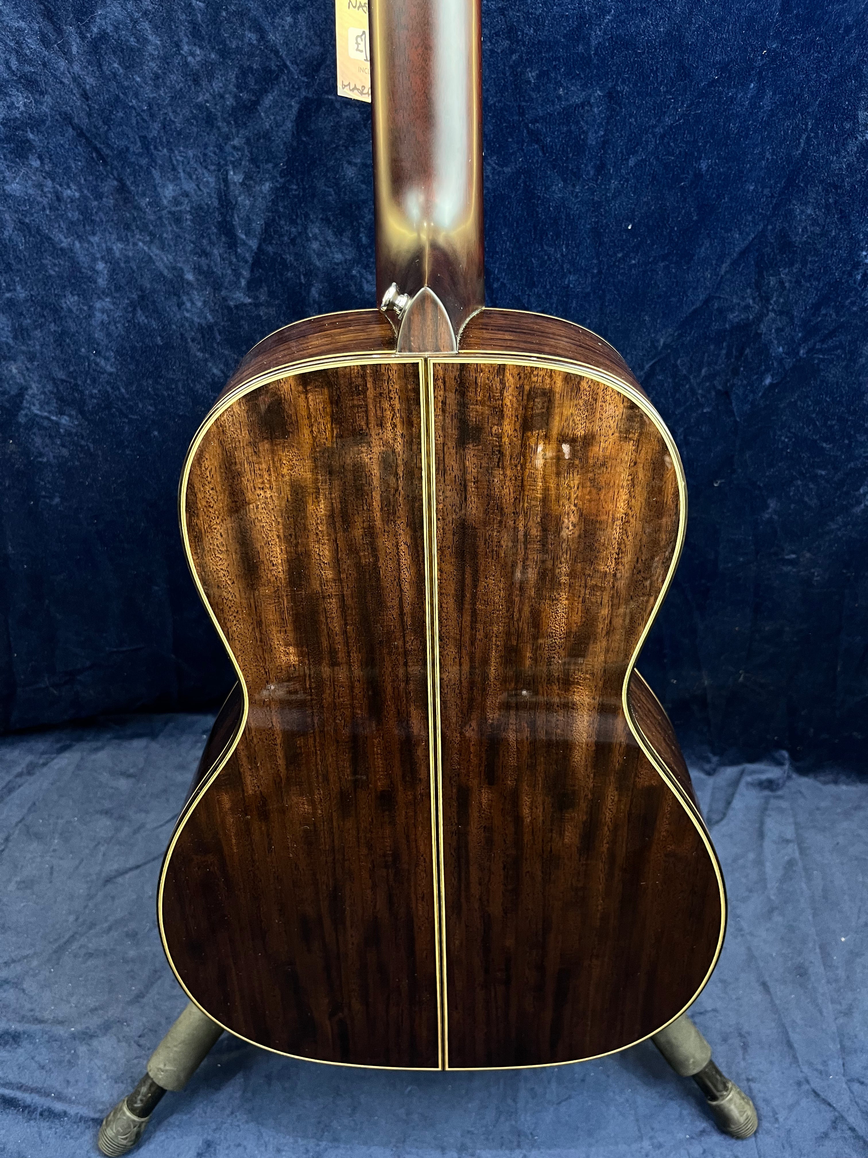 Back view of an acoustic guitar with dark, glossy wood and a central decorative strip, resting on a stand against a dark blue background.