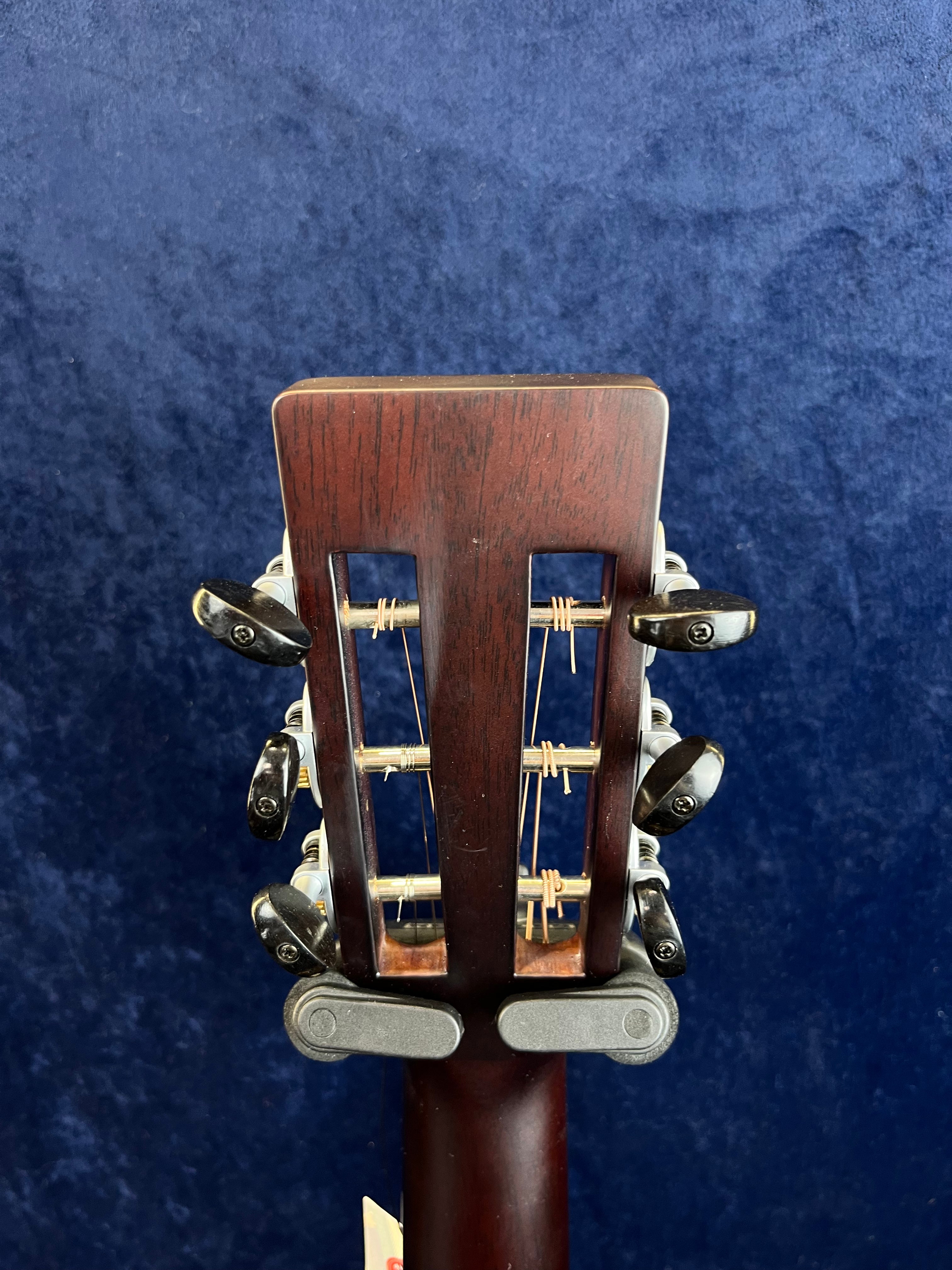 Rear view of an acoustic guitar headstock with six tuning pegs and slotted design, set against a dark blue textured background.