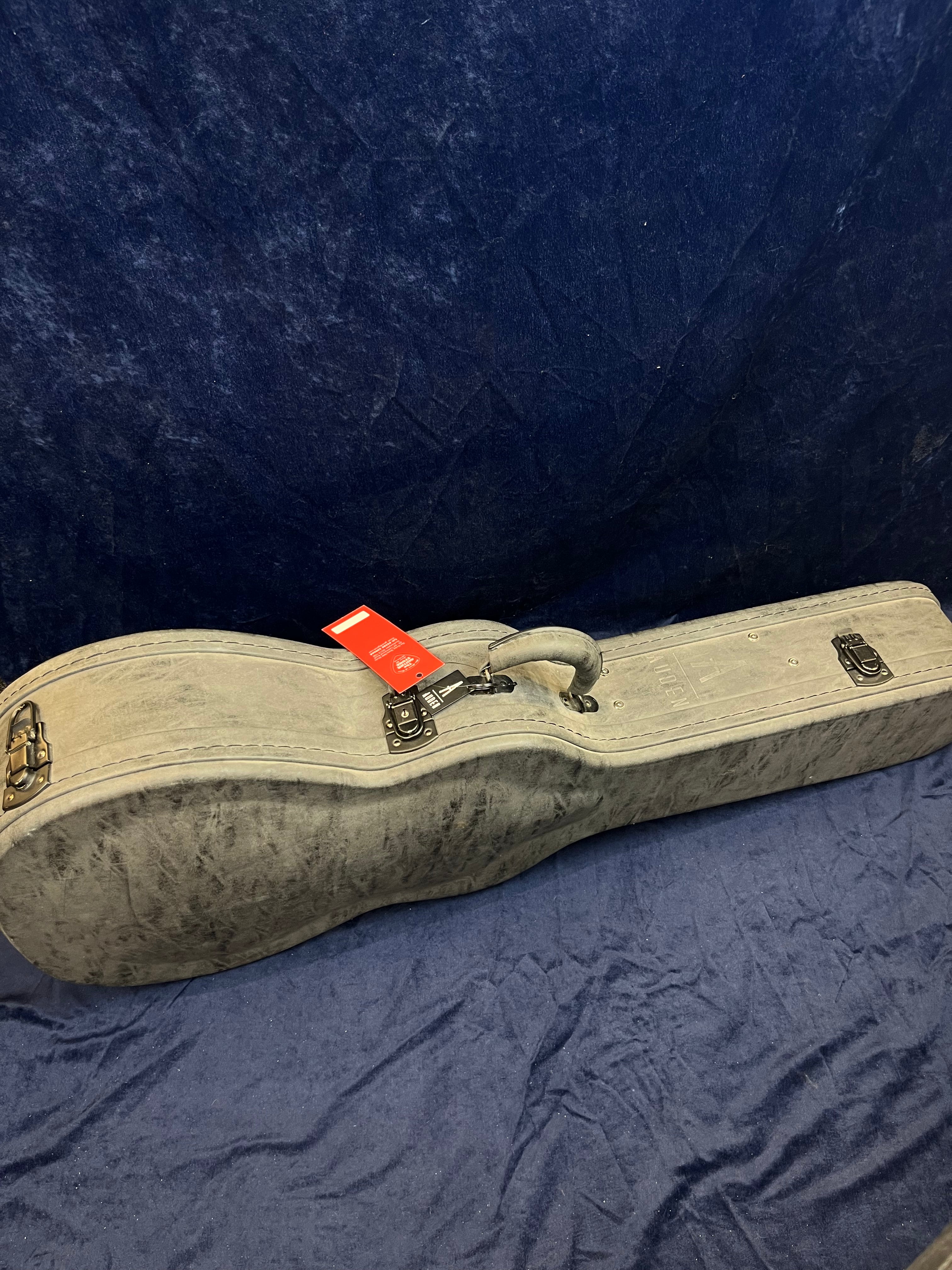 A gray hard acoustic guitar case with a handle, metal latches, and a red tag attached, resting on a dark blue fabric surface.
