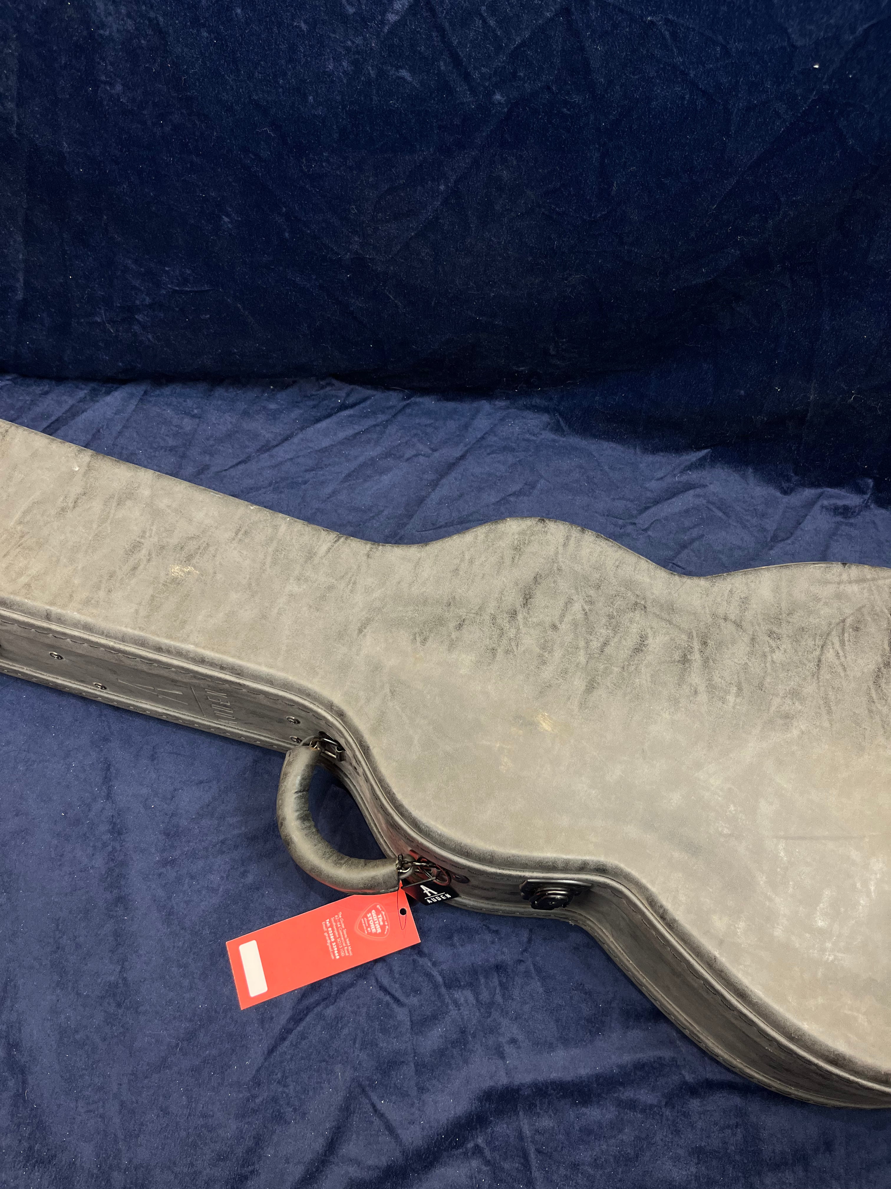 Worn, hard-shell acoustic guitar case with a handle and attached red tag, resting on a dark blue cloth background.