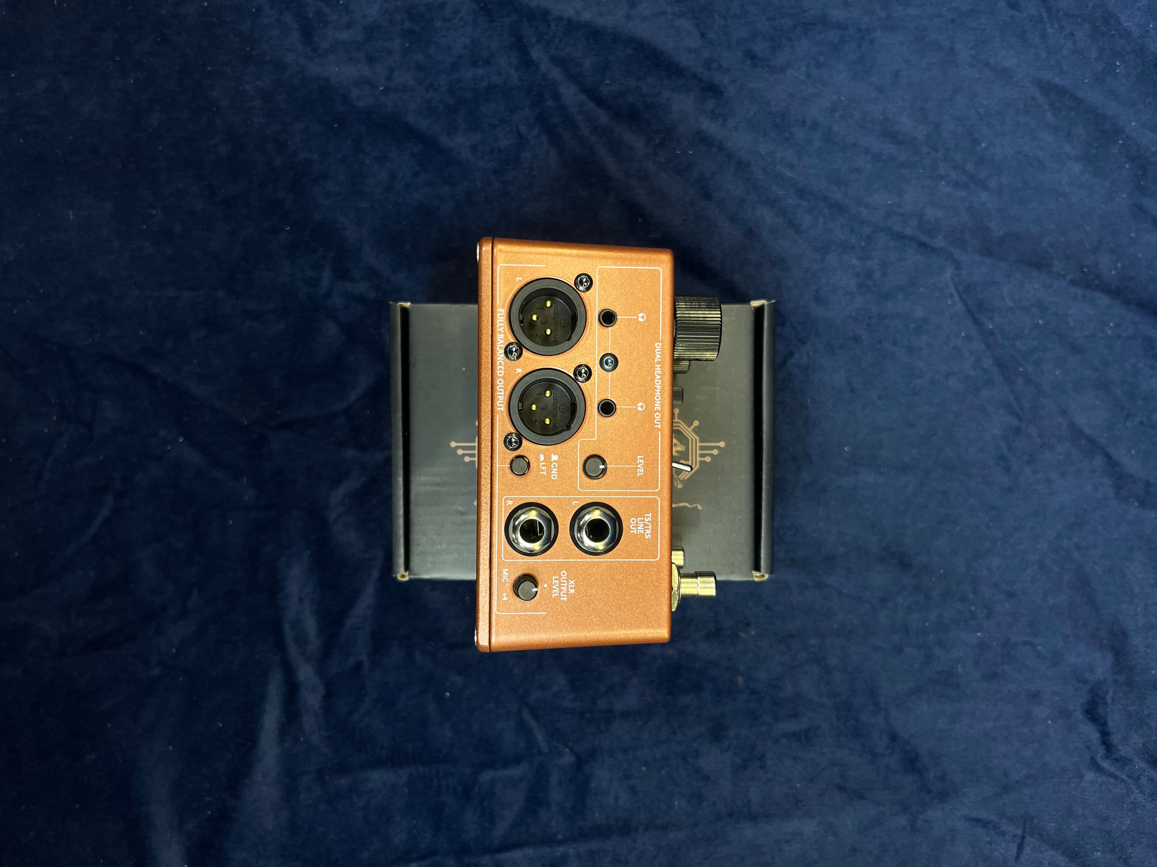 Copper-colored FX pedal with two XLR outputs, two quarter-inch outputs, control knob, switches, and labels, resting on a box against a dark fabric background.