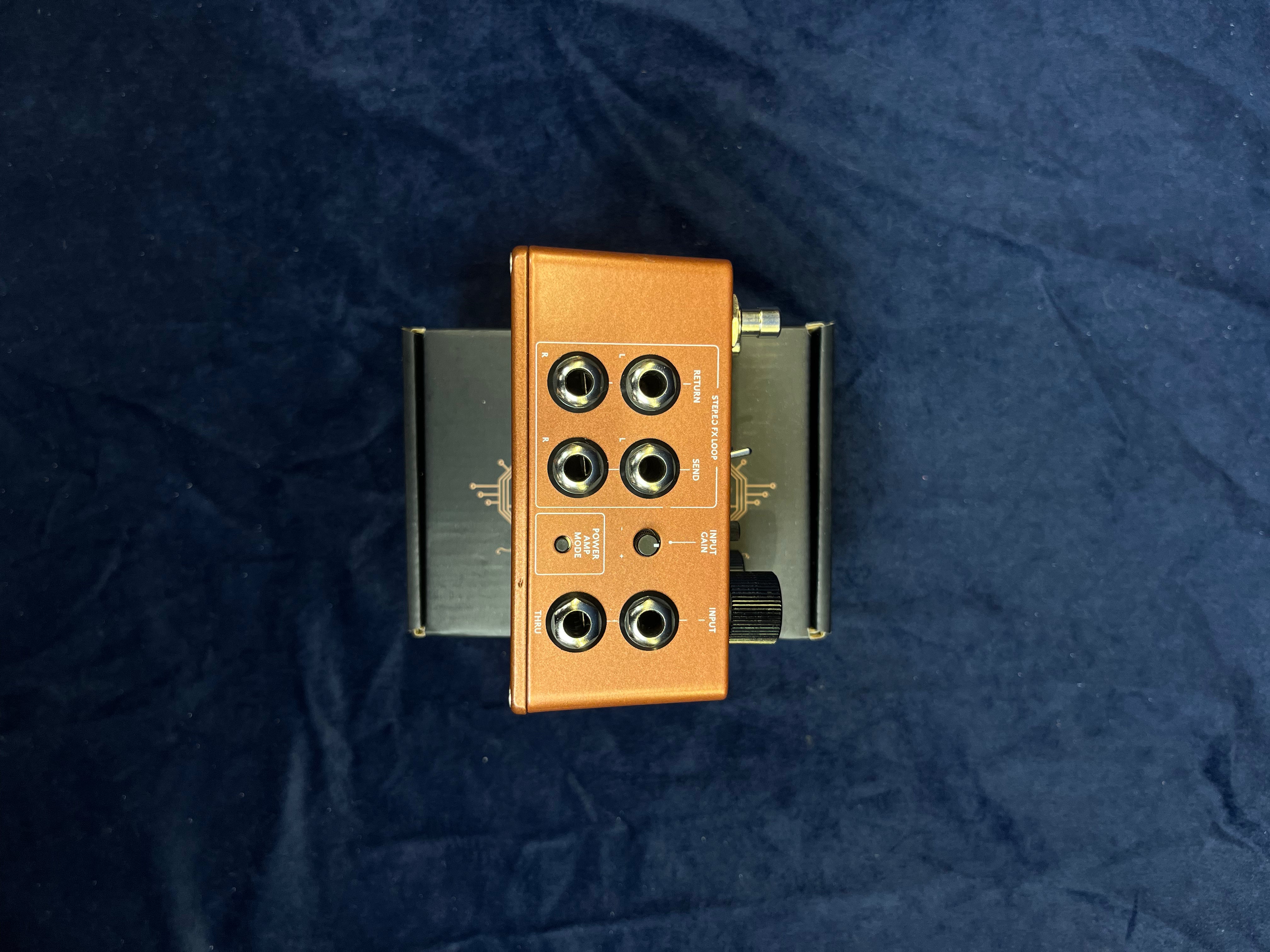Copper-colored FX pedal with six audio jacks, a power input, and black knob, placed on a black box against a dark blue background.