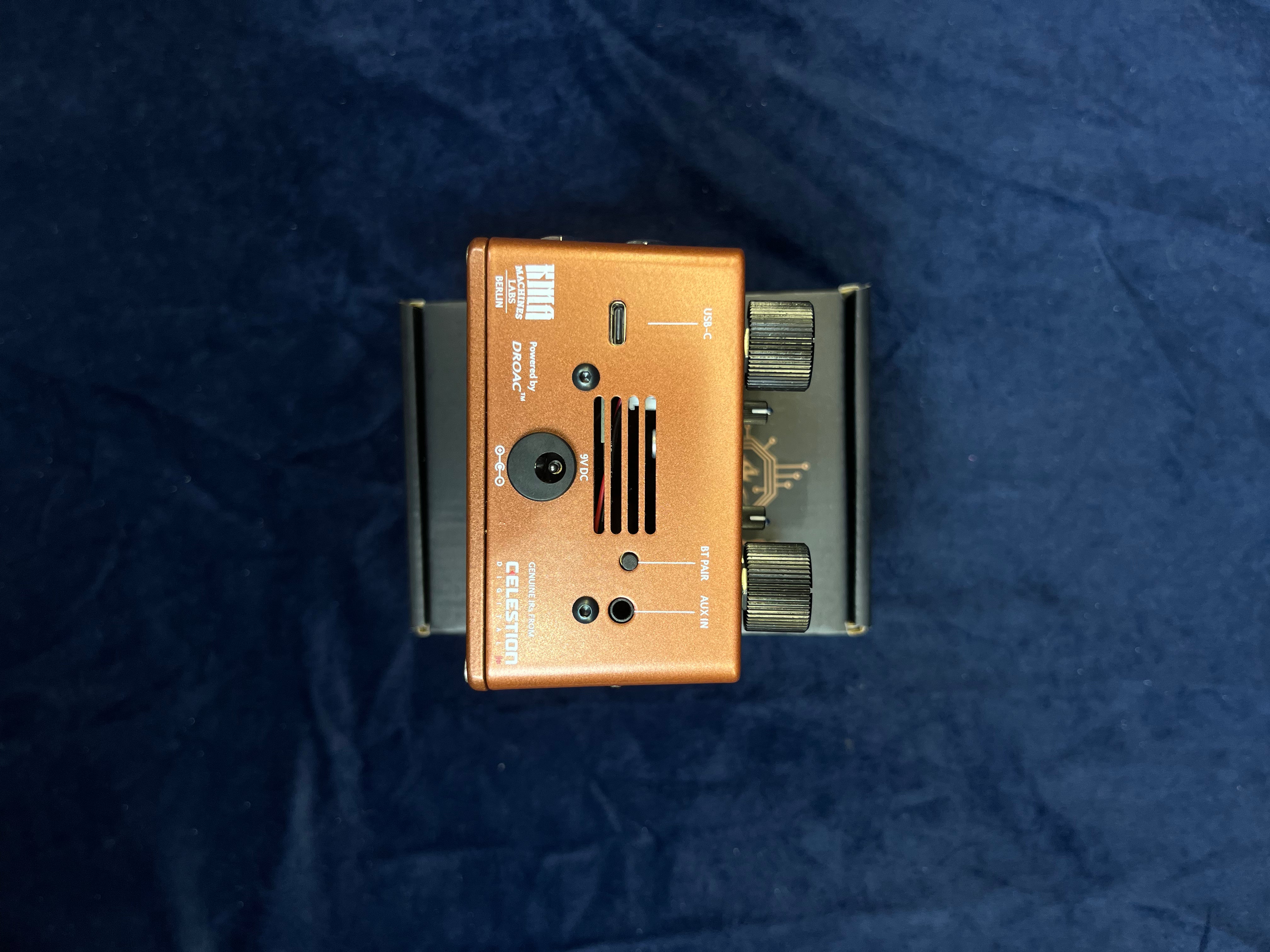 Copper colored FX pedal with two black knobs, input/output jacks, and a vented grille on top, placed on a box against a dark fabric background.