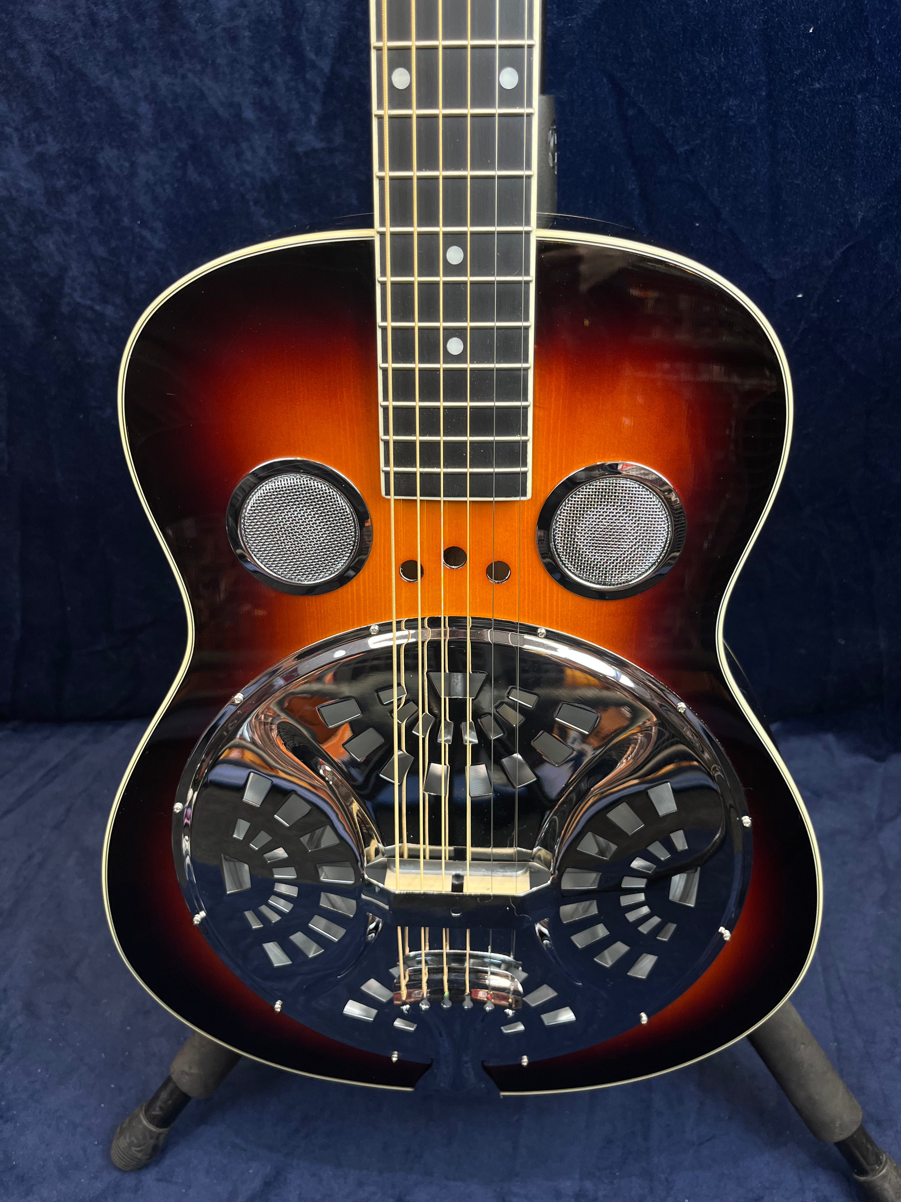 A resonator guitar with a sunburst finish, two circular sound holes, and a large chrome resonator coverplate with geometric cutouts on the body.