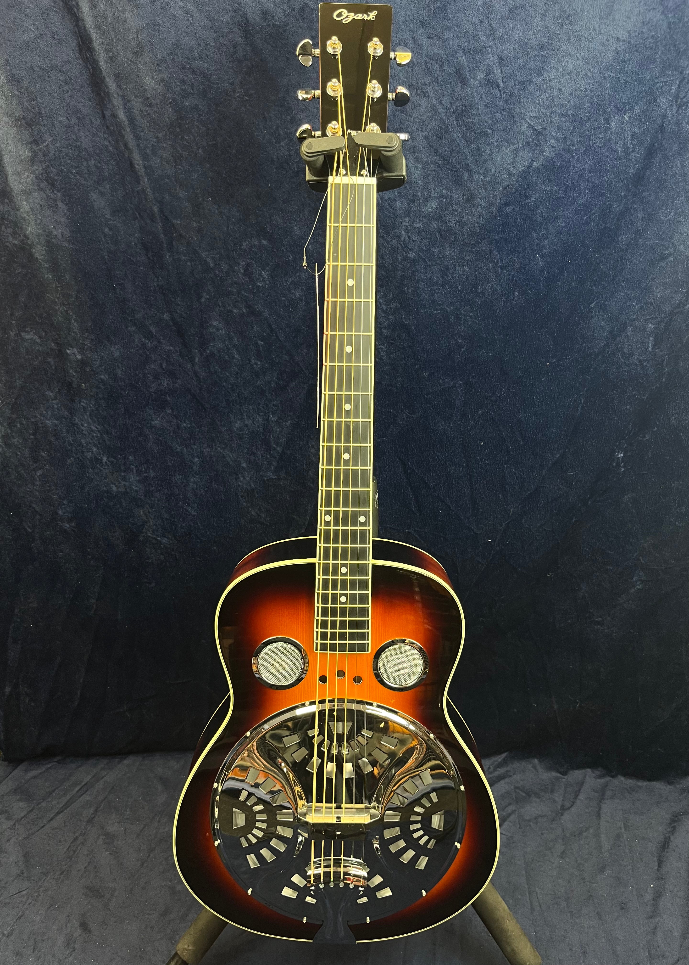 Ozark resonator guitar with a sunburst finish, chrome cover plate, two sound holes, and six strings on a dark background.