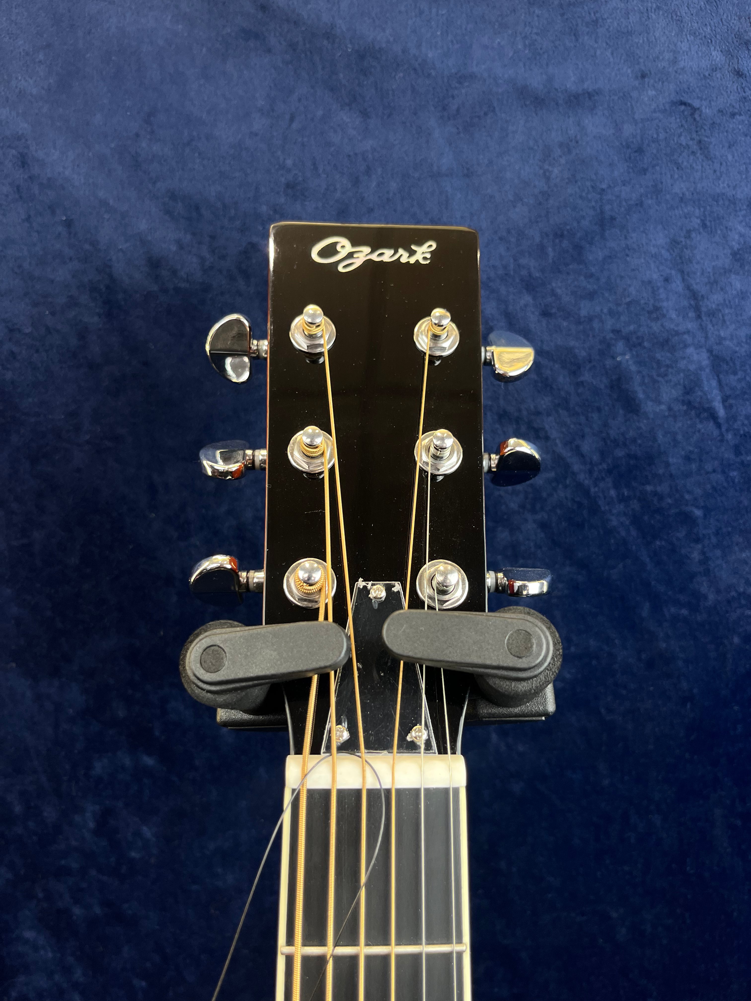 Black Ozark resonator guitar headstock with six tuning pegs, gold strings, and two black capos clamped just above the nut. Blue background.