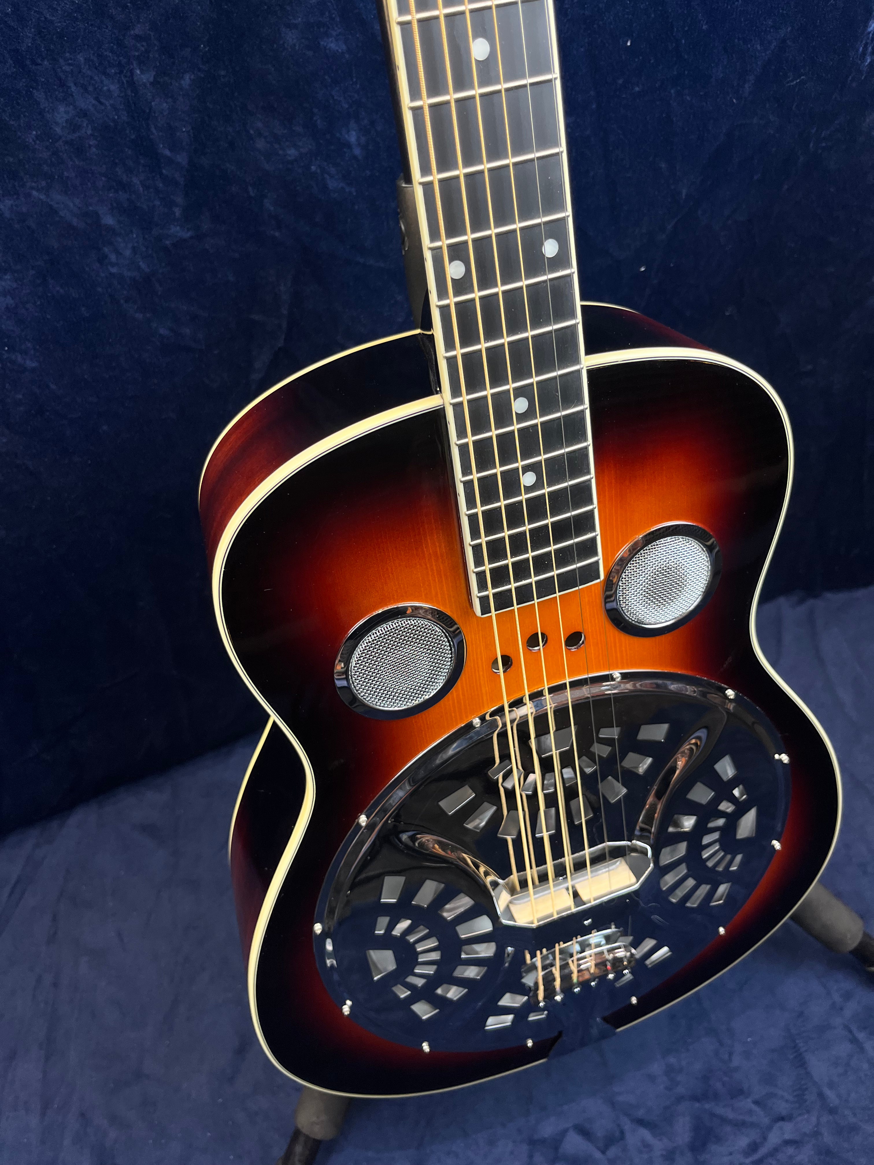 Sunburst resonator guitar with chrome cover plate, circular metal sound holes, and dot inlays on a dark wood fretboard, standing on a soft surface.