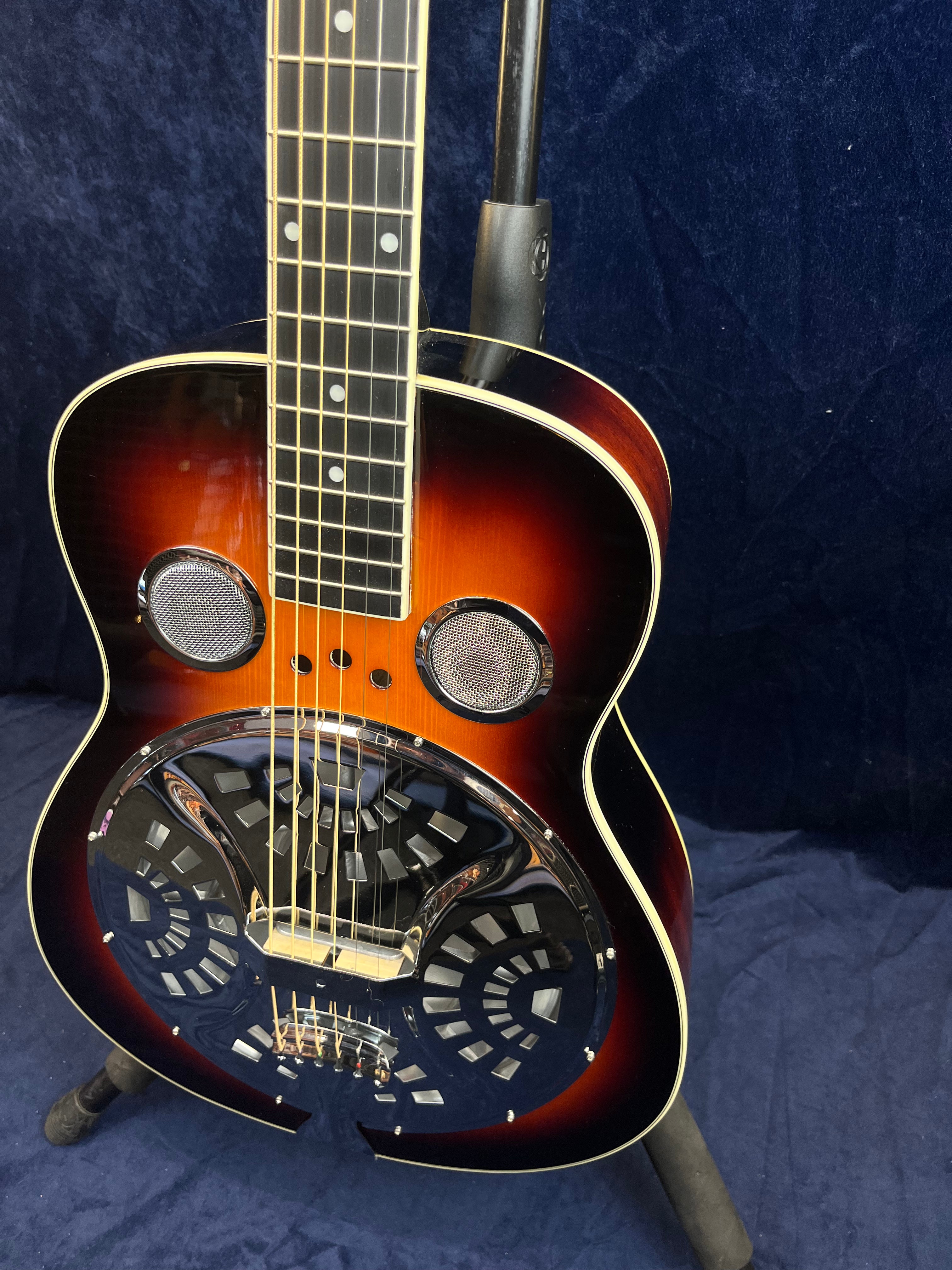 Sunburst resonator guitar with metal resonator cone and two circular screened sound holes on a dark blue background.