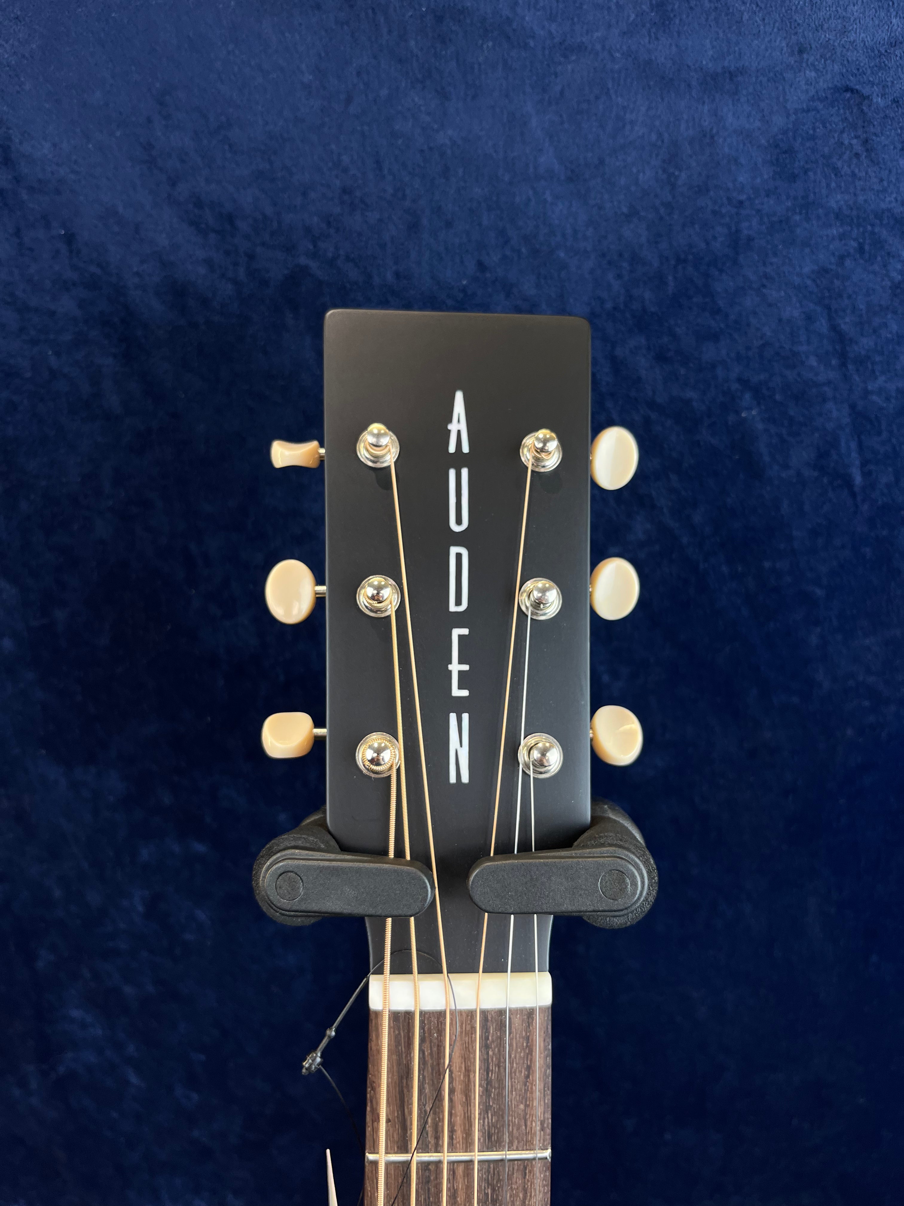 Tête d'une guitare acoustique noire avec le mot AUDEN en blanc, six mécaniques crème disposées trois de chaque côté.