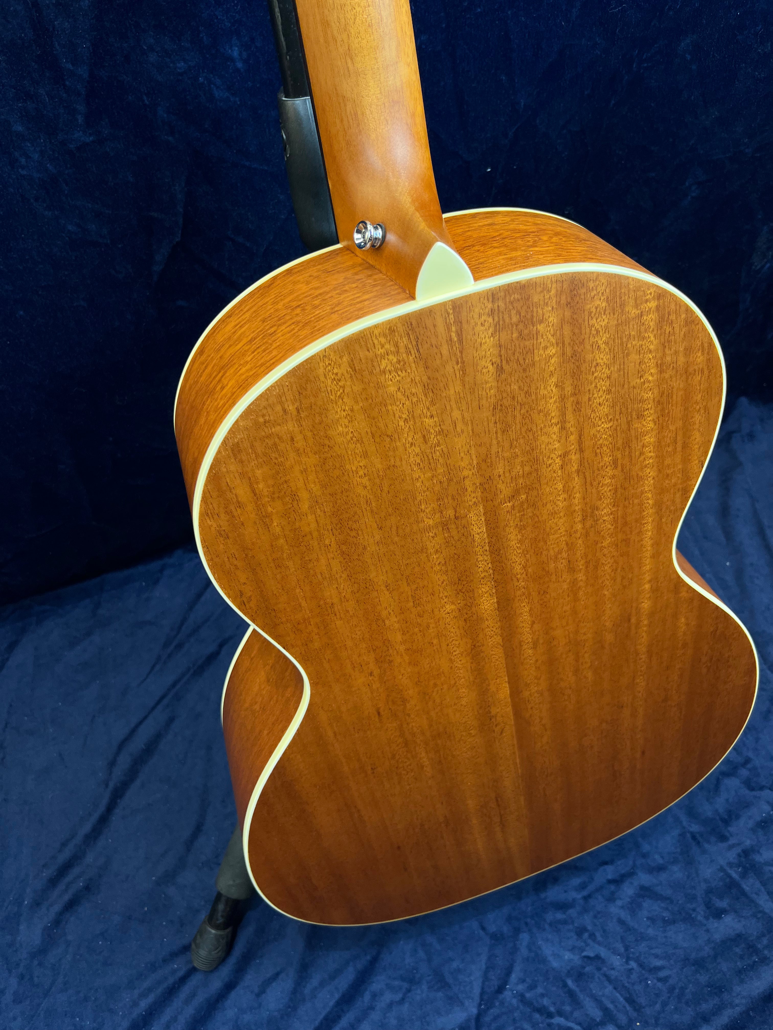 Guitare acoustique vue de dos, bois brun verni à bordure claire, fond bombé, posée sur un support, fond bleu foncé.