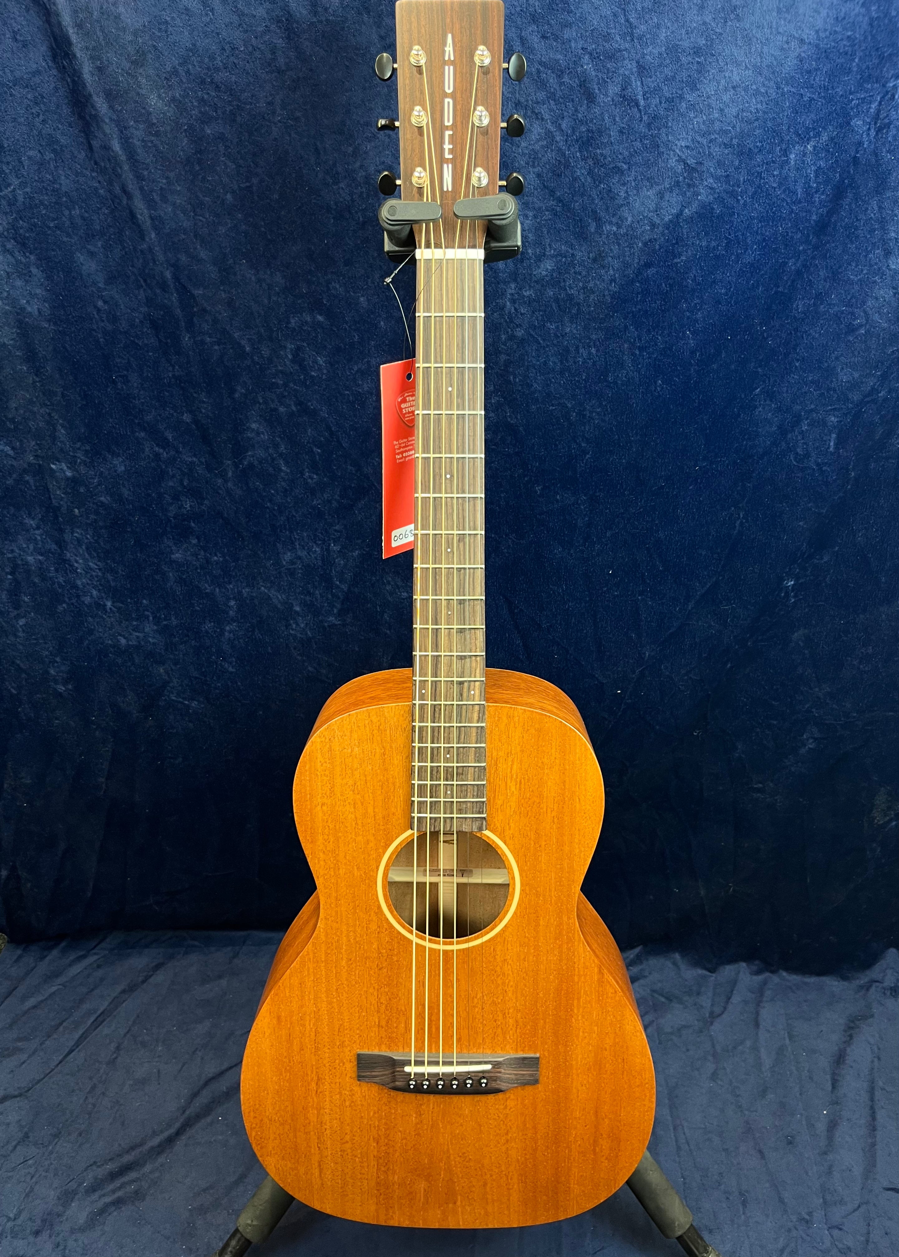 Small-bodied acoustic guitar with a natural wood finish, six strings, and a rectangular headstock marked "AUDEN," displayed upright on a stand.