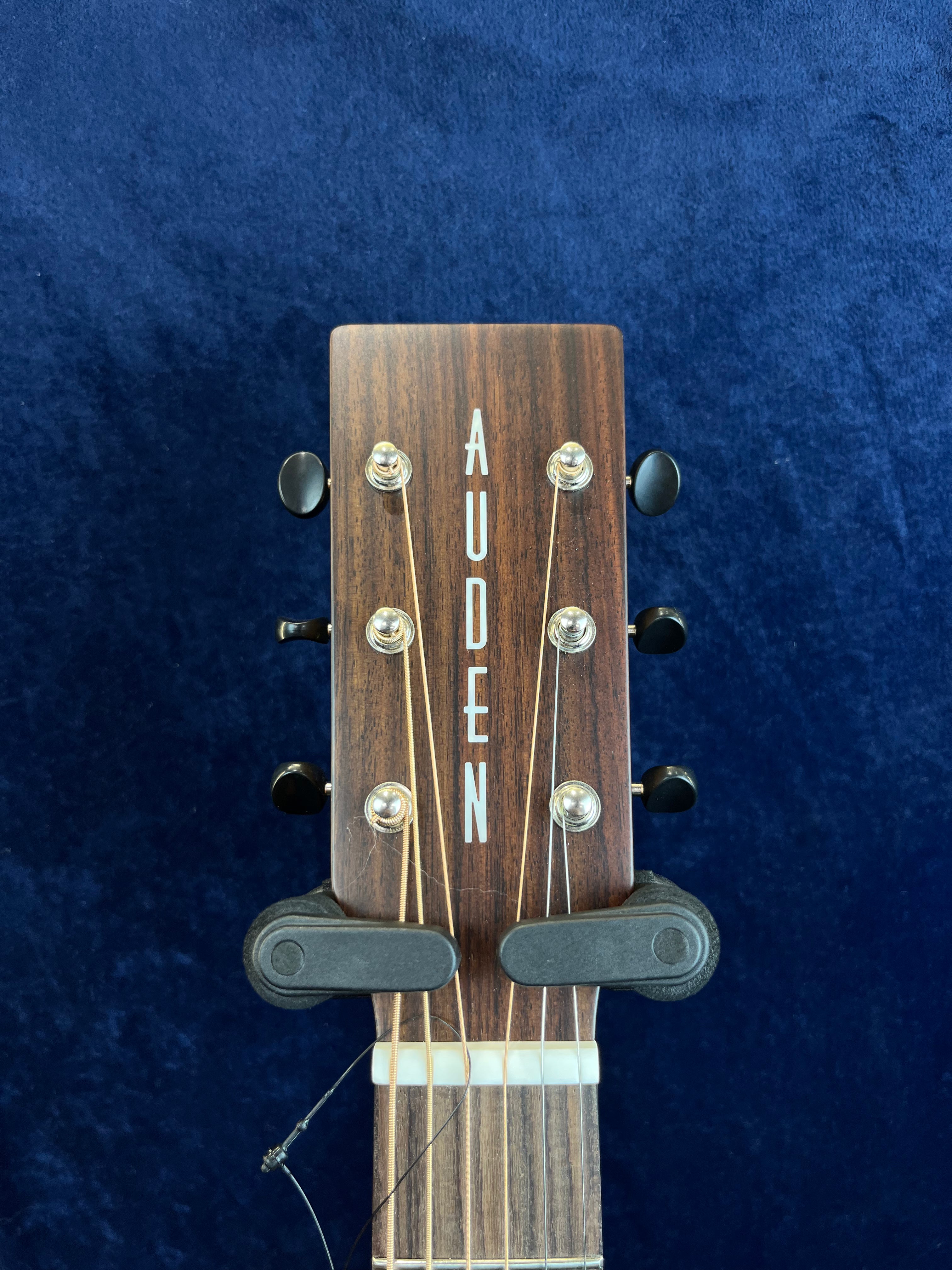 The headstock of an Auden acoustic guitar with six tuning pegs, strings attached, and a dark wood finish against a dark blue background.