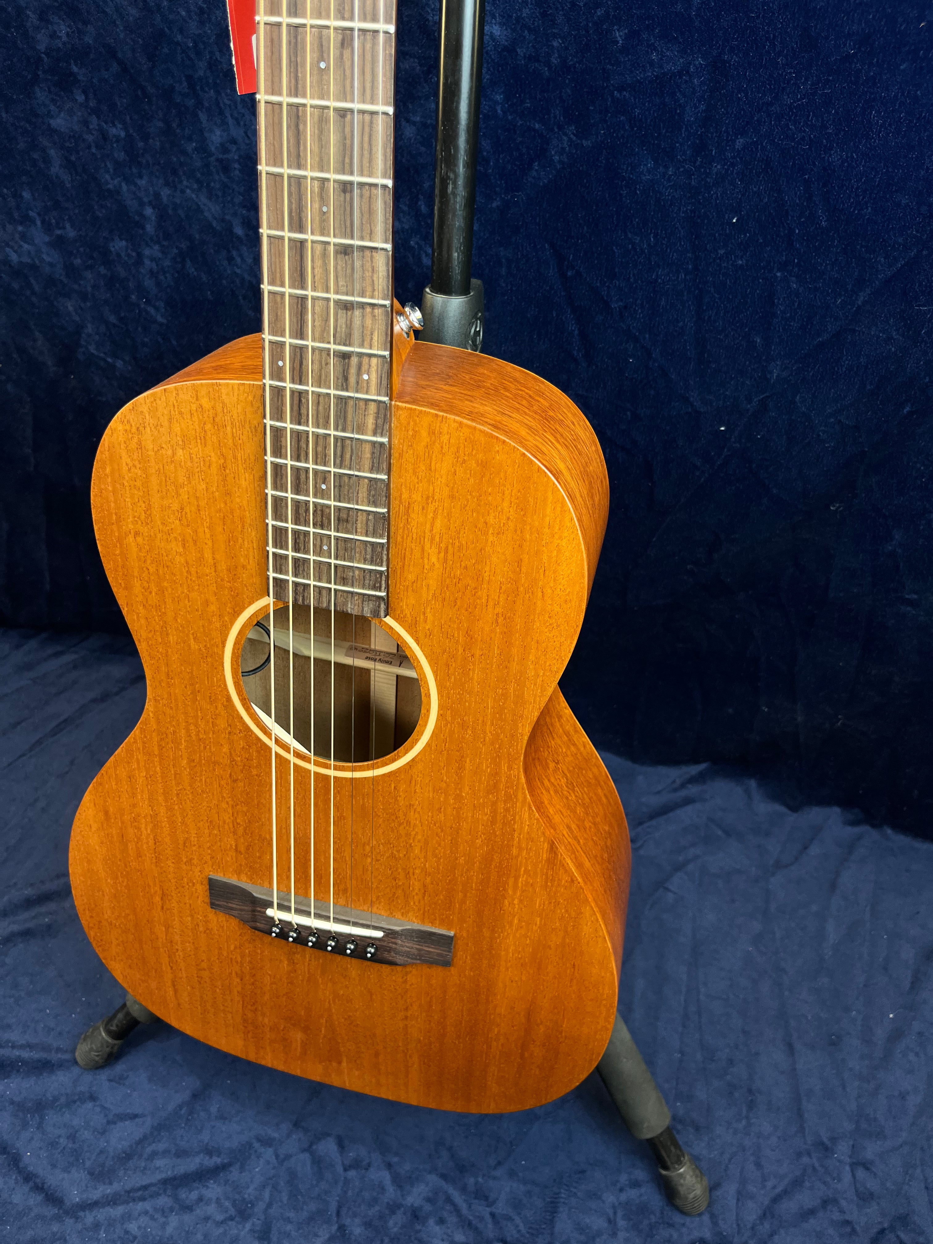 Small-bodied acoustic guitar with a natural wood finish, six strings, and a dark wood fretboard, resting on a stand against a dark blue background.